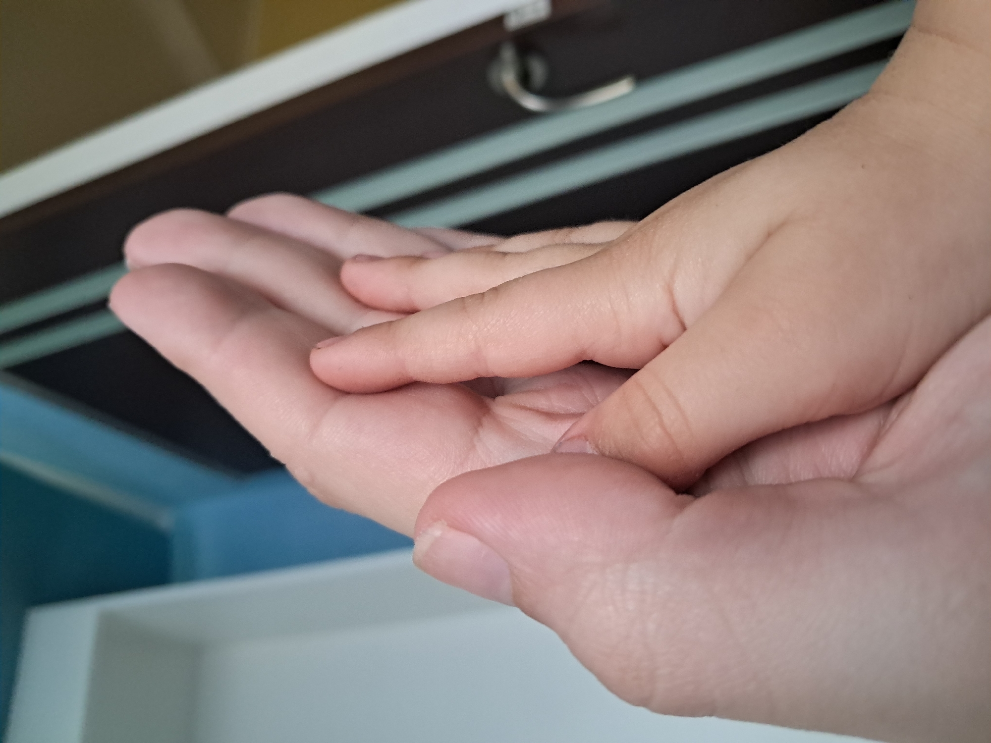 The image is a close-up, eye-level shot of two hands, one adult and one child, clasped together. The adult's hand, on the left, is larger and shows visible knuckles and palm lines. The child's hand, on the right, is smaller and smoother, with fingers resting on the adult's palm. The background is blurred, but appears to be an indoor setting with a door, possibly in a home or apartment. The lighting is soft, highlighting the textures of the skin. The exact location in the city of Unknown, Unknown is not discernible from the image.