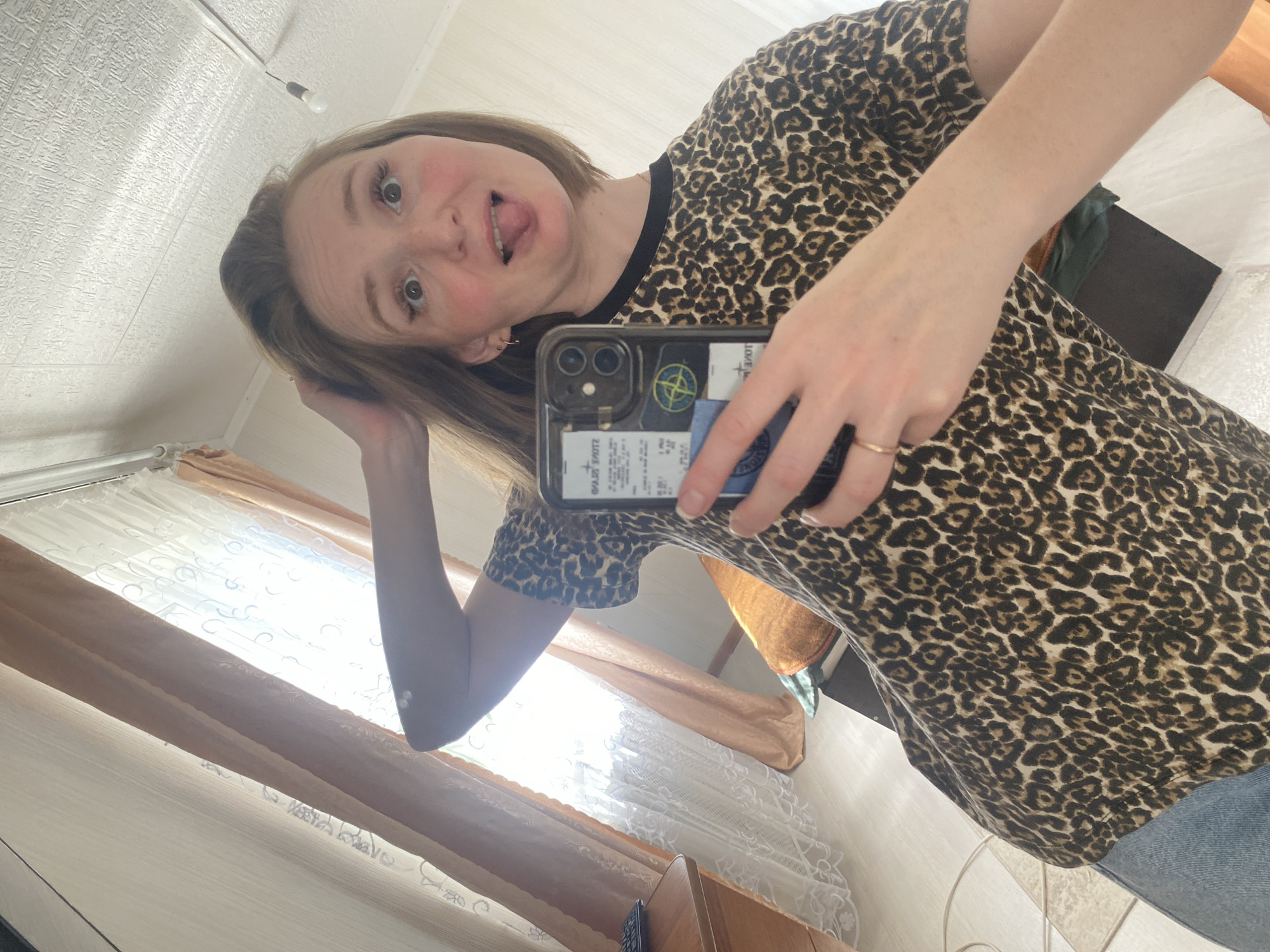 A young woman with reddish-brown hair is taking a selfie in a room. She is wearing a leopard-print t-shirt and blue jeans. Her left arm is raised, touching her hair, and her right hand is holding a phone. She is sticking her tongue out at the camera. The background shows a window with curtains, a desk with a keyboard, a pillow, and a light fixture on the ceiling. The scene appears to be an interior residential setting, likely an apartment. The specific location in Chaikovsky, Russia, is not discernible from the image itself.