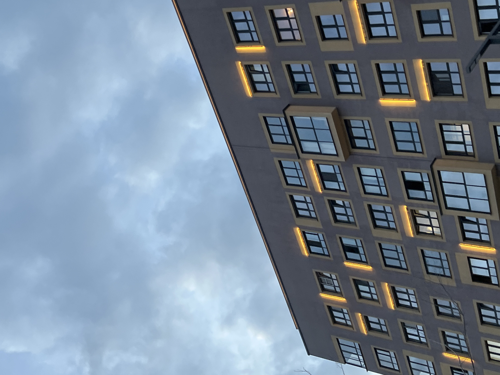 This image shows the exterior of a modern apartment building in Yekaterinburg, Russia, under a cloudy sky. The building is constructed with a grey facade and features numerous windows arranged in a grid pattern. Many of the windows are illuminated from within, casting a warm yellow glow. Some windows are open, and the building has a distinctive architectural element with projecting window boxes. The lighting suggests it is either early morning or late evening. No people are visible in the frame.