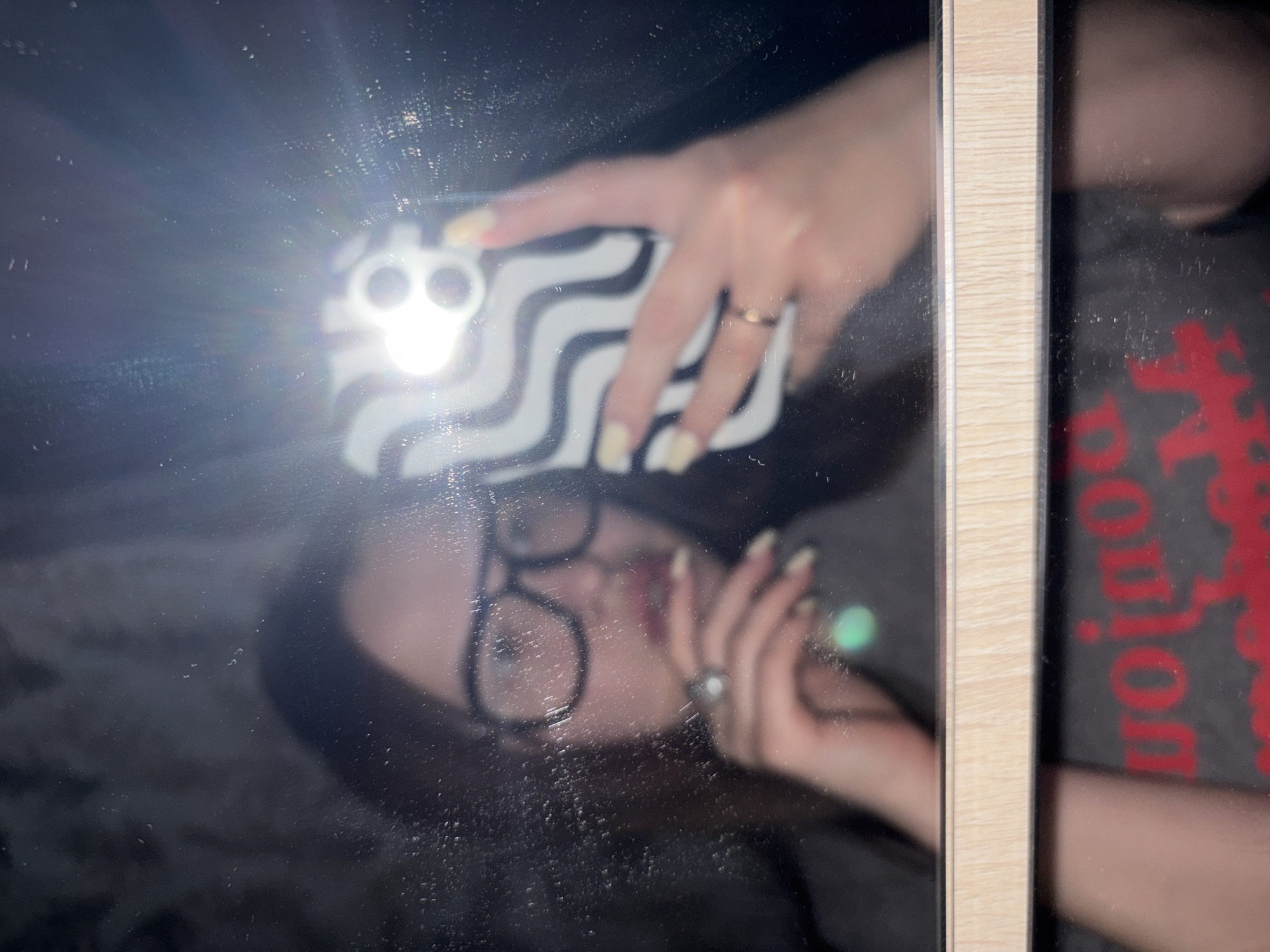 The image shows a blurry reflection in a mirror, partially obscured by water spots. A person is taking a selfie with a phone, its zebra-striped case and illuminated flash visible. The person wears glasses and rings. A reflection of another person wearing a grey t-shirt with red lettering is also visible on the right side of the mirror. The mirror is framed in light wood. The location and context of the image are unidentifiable, making it impossible to link to any location in the city of Port-Katon, Russia, or to make forensic descriptions about it.