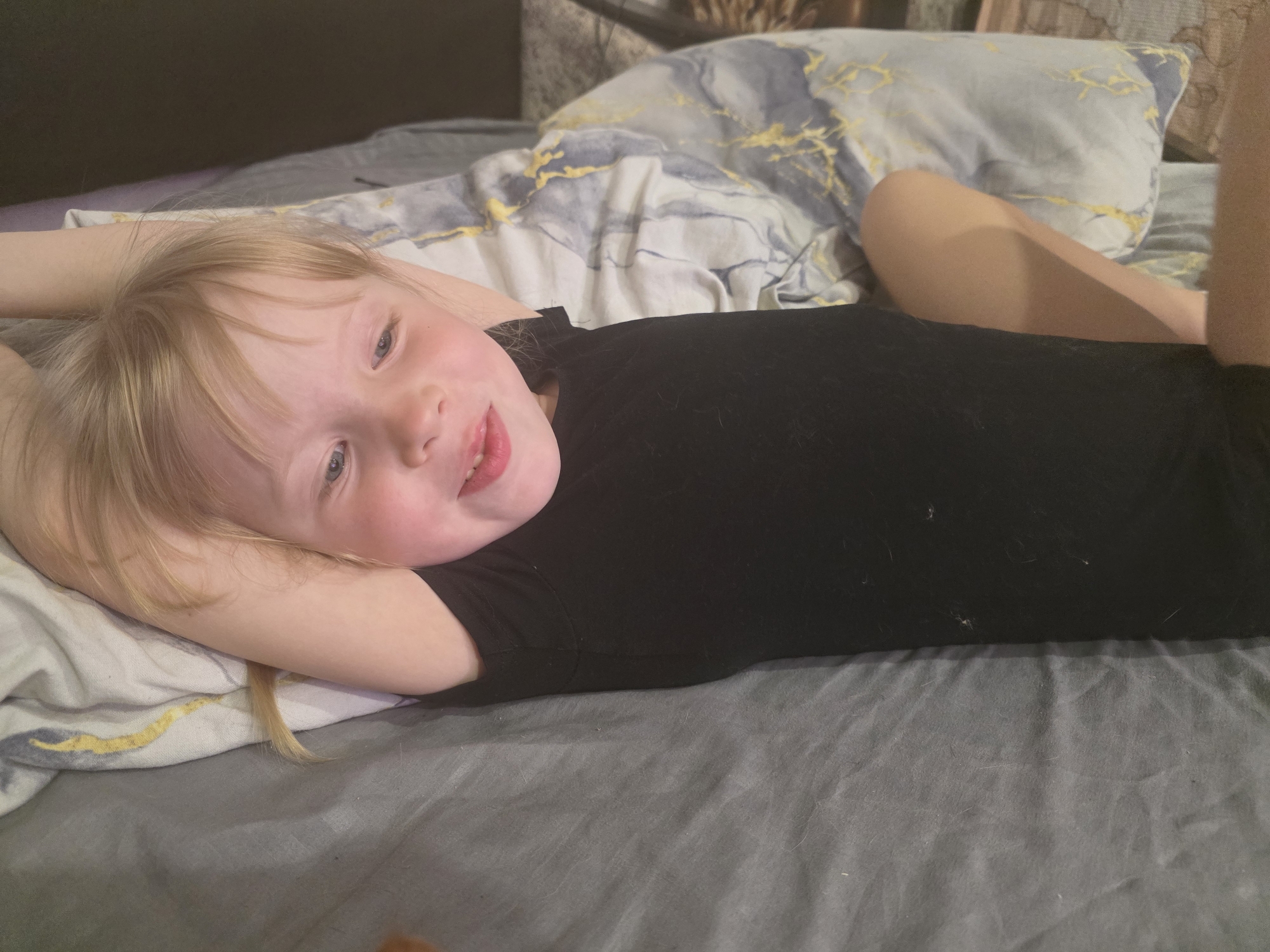 A young child with blonde hair is lying on a bed. The child is wearing a black shirt. The bed has grey sheets and a pillow with a blue and gold design. The location is not discernible from the image.