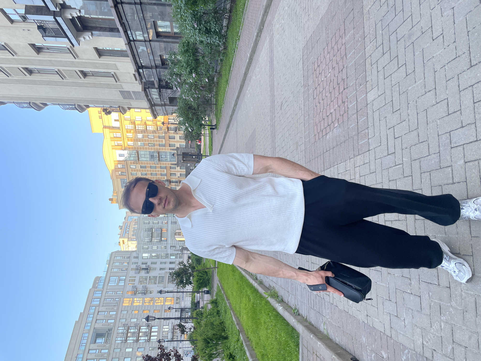 A male, appearing Caucasian, with short light brown hair and sunglasses, stands on a paved pedestrian path in an urban environment in Saint Petersburg, Russia. He wears a white, short-sleeved, ribbed knit polo shirt with an open V-neck collar, loose-fitting black trousers, and white and silver athletic shoes. His left hand holds a small black zippered bag with a strap, while his right hand is positioned within his trouser pocket. Visible veins are present on his left forearm.
The path is constructed from interlocking pavers, primarily light grey in a herringbone pattern in the foreground, transitioning to sections with red-brown pavers further along. Green grassy areas run parallel to the path on both sides, interspersed with small bushes, some displaying light-colored blossoms. Multi-story residential buildings, predominantly light-colored (beige or off-white), line both sides of the path. These buildings feature numerous windows and balconies with metal railings. A sign displaying "САЛОН КРАСОТЫ" (Beauty Salon in Russian) is visible on a building on the left. Windows on buildings in the background reflect warm, golden sunlight. The lower section of the building on the right is clad in a dark, possibly black, stone or tile material. Multiple dark street lamps are positioned along the pathway. The sky above is clear and bright blue. The lighting suggests natural daylight, likely late afternoon or early evening due to the direction and warmth of reflected sunlight.