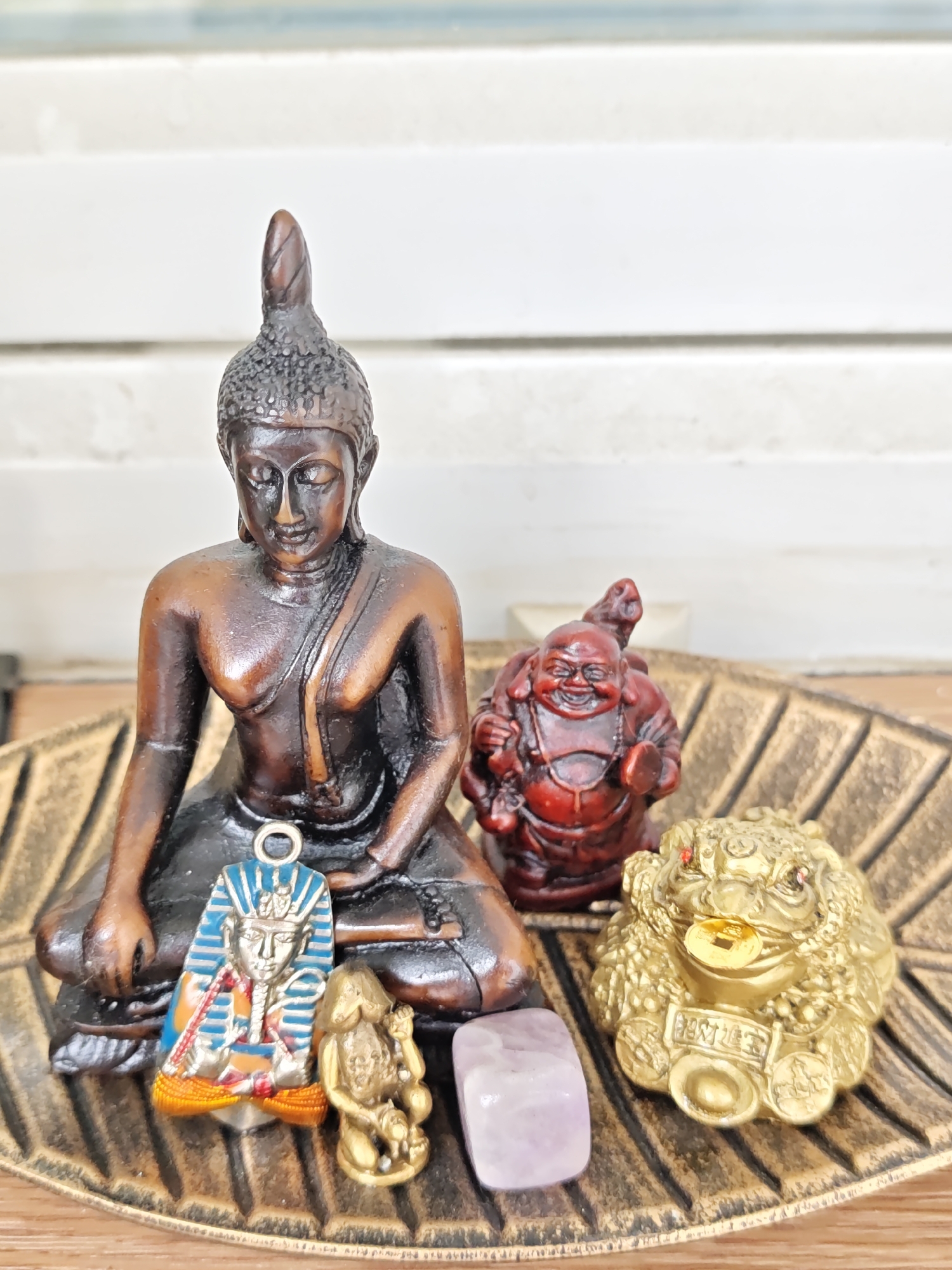 This image captures a collection of decorative figurines arranged on a textured golden tray, which rests on a wooden surface. The figurines include a seated bronze Buddha statue, a small red figurine of a jovial man holding a bag, a gold-colored frog with a coin in its mouth, a small gold figurine of a monkey holding a peach, a pendant featuring an Egyptian pharaoh's face, and a pale purple crystal. The background is a blurred white surface with horizontal lines, suggesting it could be a wall or a piece of furniture. The lighting appears to be natural, casting soft shadows. No people are visible in the image. The scene does not depict any specific location in Saint Petersburg, Russia.