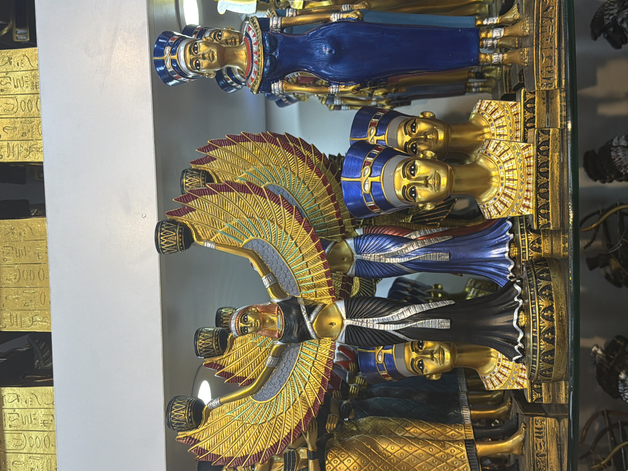 This is a frontal, eye-level medium shot of a display of Egyptian-themed souvenirs, likely in a shop in Sharm El Sheikh, Egypt. The foreground features several golden statues and busts. Prominently in the center is a winged statue of a goddess, possibly Isis, with outstretched golden wings adorned with red and blue accents. She wears a black and white garment and holds two cylindrical objects above her head. To her right, a tall, slender female statue in a vibrant blue dress stands with her hands at her sides. To the left of the central winged statue are several golden busts of pharaohs wearing traditional nemes headdresses, two of which are clearly visible. Behind these figures, more golden statues and headdresses are partially visible. The background consists of a white shelf with rows of golden rectangular plaques inscribed with hieroglyphs. The overall impression is a collection of ornamental items intended for tourists, showcasing Egyptian art and mythology.