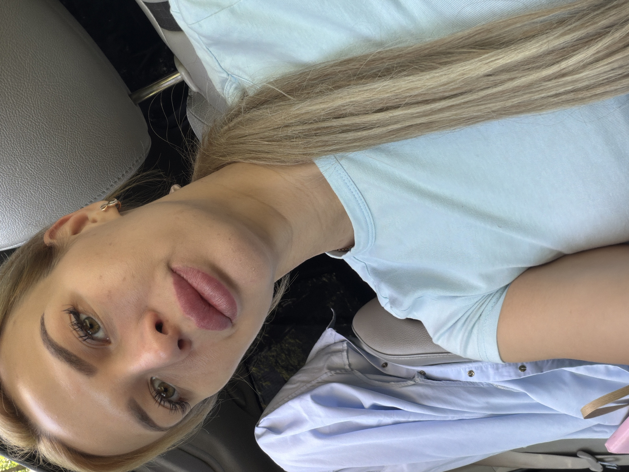 A young woman with blonde hair pulled back in a ponytail is shown in a close-up selfie. She is wearing a light blue t-shirt and a light blue button-down shirt is visible draped over a car seat next to her. A pink purse is also visible in the lower left corner. The scene appears to be inside a vehicle, possibly a car, with the gray leather headrest of a car seat visible in the background. The lighting suggests it is daytime. The location is specified as Kyiv, Ukraine.