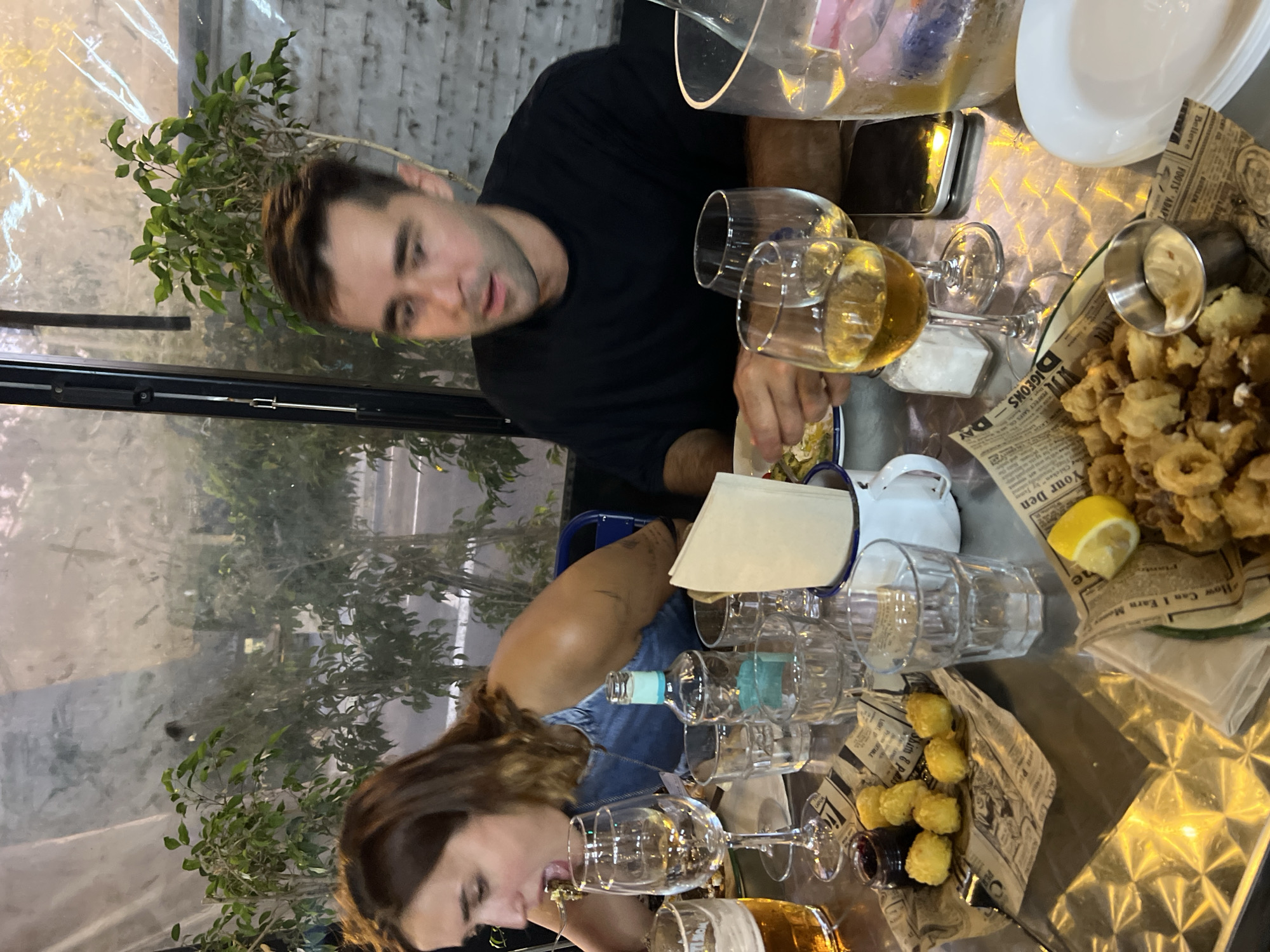 The image depicts a dimly lit outdoor dining scene, likely at a restaurant in Barcelona, Spain. Two individuals, a woman and a man, are seated at a metallic table laden with food and drinks. The woman, on the left, is leaning forward with her mouth open, in the process of eating. She has brown hair and is wearing a denim top. The man, on the right, is looking to his left with his mouth slightly agape, appearing to be mid-conversation or react to something. He has dark hair and is wearing a black t-shirt.
On the table, there are several glasses, including wine glasses containing a golden liquid, and a beer glass. A bottle of clear liquid with a light blue label, possibly water or a spirit, stands prominently. Appetizers are arranged on newspaper-like paper, including a small dish of dark jam with several small, fried spherical items, and a larger serving of fried seafood, possibly calamari, with a small metal cup of dipping sauce. A lemon wedge is also visible. The background features greenery, suggesting a plant-filled outdoor space, and a glimpse of a street with streetlights and a building. The overall ambiance is casual and convivial.