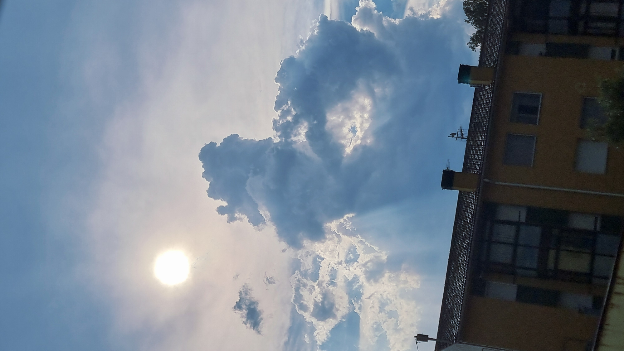 The image shows a building with a yellow facade and a tiled roof under a partly cloudy sky. The sun is visible in the upper left portion of the sky, partially obscured by clouds. Dramatic cloud formations are present in the center, with sunbeams piercing through them. The building appears to be residential, with several windows visible. The location is specified as Sant Joan Les Fonts, Spain.
