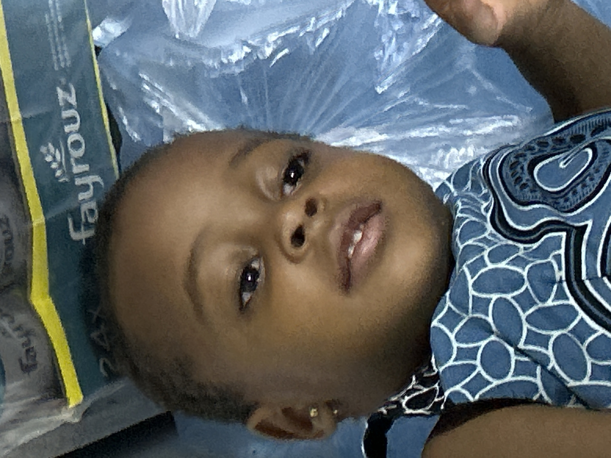 A young child lies on a blue surface, possibly covered with plastic sheeting. The child is wearing a blue and white patterned shirt and has a gold stud earring in their left ear. To the left of the child is a green and yellow package with the word "Fayrouz" visible. The location is given as Ihiagwa, Nigeria, but there is no visual indication of this in the image.