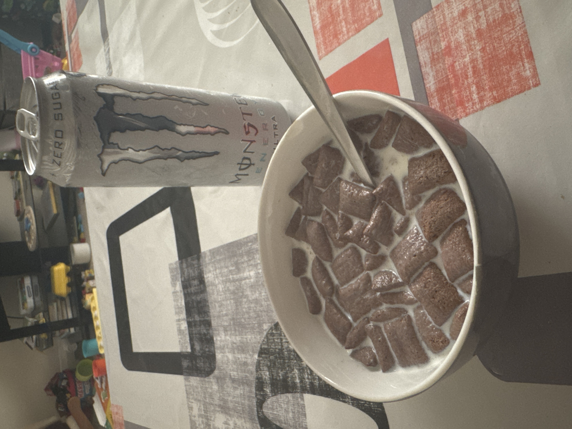 A can of Monster Energy Ultra Zero Sugar drink lies horizontally beside a bowl of chocolate cereal with milk and a spoon on a tabletop with a patterned tablecloth. The photograph appears to have been taken indoors. There is no information to connect this image to Welshpool, United Kingdom. No people are visible in the image.