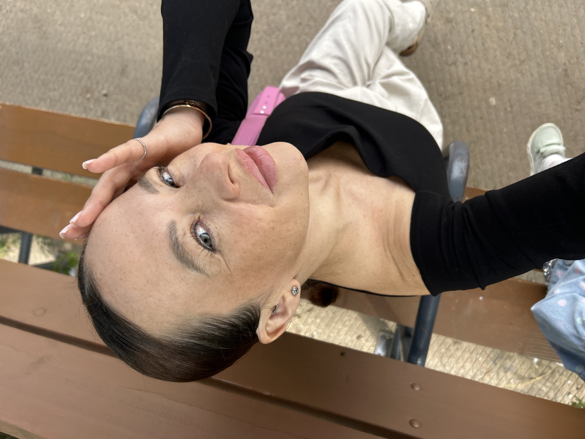 A woman is sitting on a park bench in Chaikovsky, Russia, taking a selfie. She has her right hand on her forehead and is looking up at the camera with a slight smile. She is wearing a black top with a white overlay and white pants. Her hair is pulled back in a ponytail. The bench is made of wood and metal, and the ground is paved with concrete. There are trees and foliage in the background.
