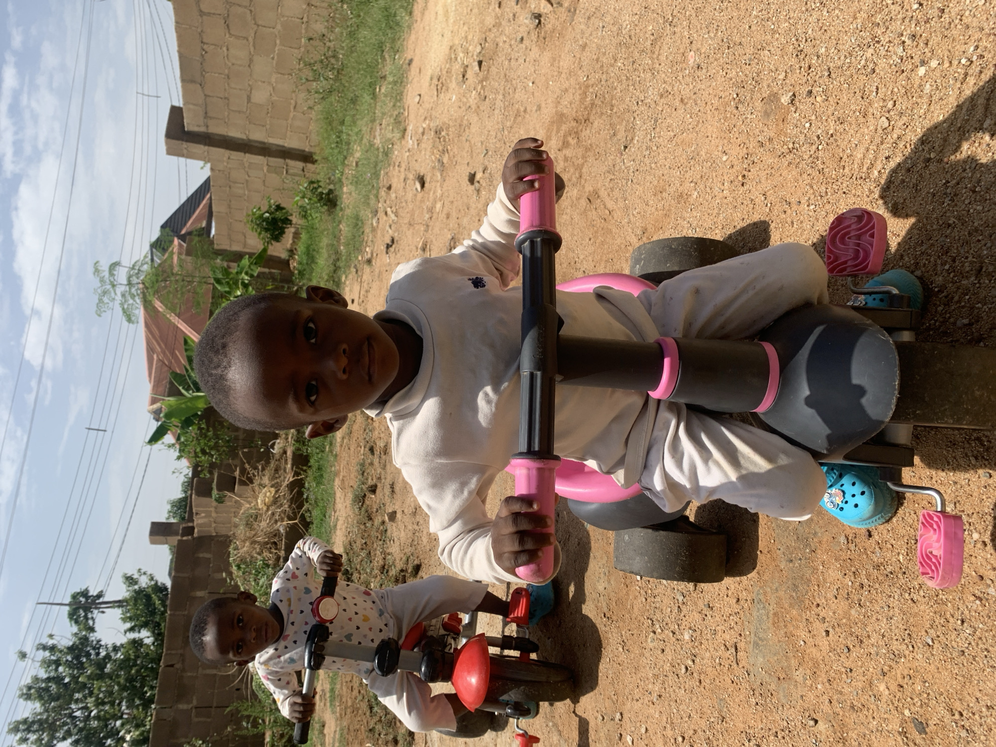 Two children are outdoors in Gura Top, Nigeria. One child, wearing a white long-sleeved shirt and pants, sits on a black and pink toy vehicle. The child looks directly at the camera and grips the pink handlebars. The other child, wearing a patterned white long-sleeved shirt and pants, is also riding a red and black toy vehicle. They are on a dirt surface. Behind them is a brick building, trees, power lines, and a partially visible house under a blue sky with clouds.