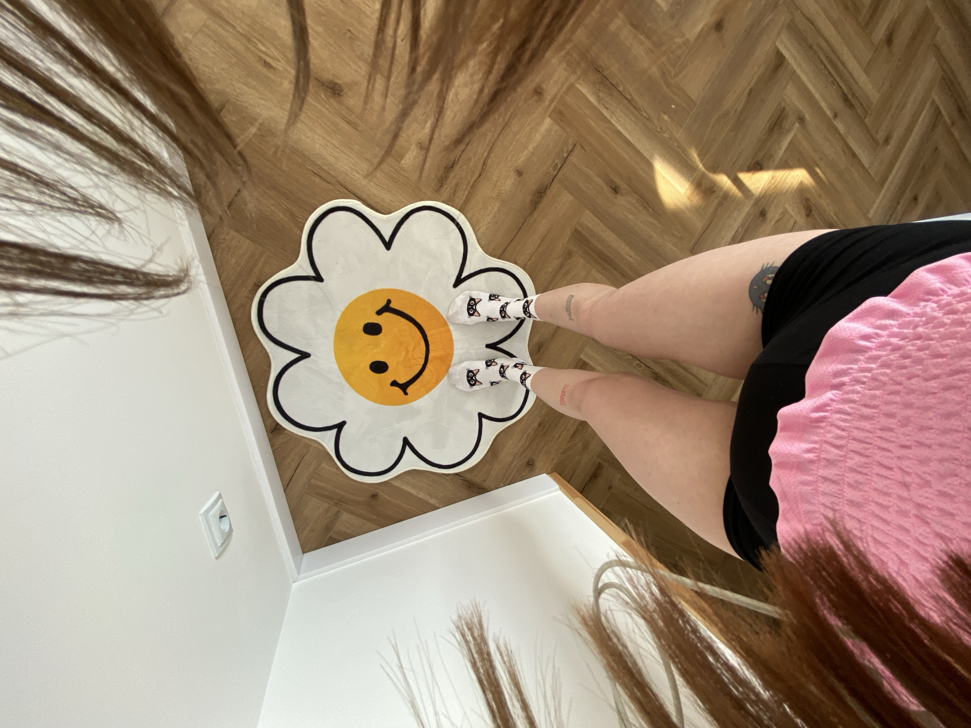 A high-angle shot shows a person standing on a wooden floor in front of a white wall with a light switch. The person is wearing a pink top, black shorts, and white socks with a black cat pattern. The person is standing on a daisy-shaped rug with a smiley face in the center. The person has brown hair. The location is unknown, and the city of Izhevsk, Russia, cannot be confirmed.
