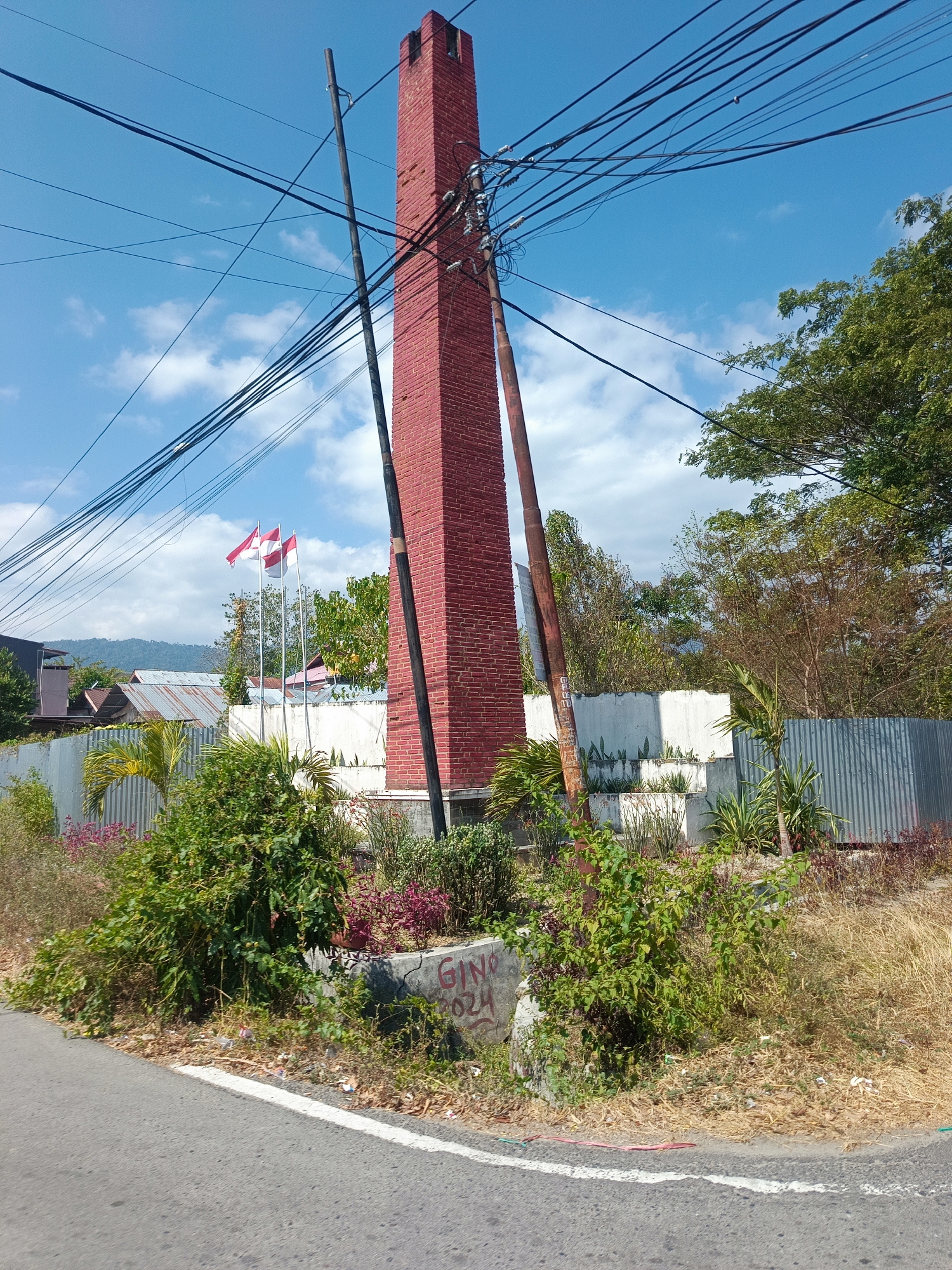 In Suwawa, Indonesia, a tall, red brick tower stands under a bright blue sky dotted with white clouds. The tower is positioned in the foreground, slightly to the right of the center of the image. It is a rectangular structure with visible brickwork and appears to be a chimney or a monument. Several utility poles and a dense network of black electrical wires are strung across the sky and around the tower, creating a complex visual.
In front of the tower, three Indonesian flags, red and white, fly from poles. These flags are partially obscured by the foliage.
The base of the tower and the surrounding area are landscaped with various plants, including green bushes, low-lying pink and purple flowers, and some dry grass. A concrete structure with graffiti, including the partial word "GIN" and numbers, is visible at the base of the tower.
Behind the tower and flags, a white wall and a corrugated metal fence demarcate the property. Beyond the fence, several buildings with corrugated metal roofs are visible, suggesting a residential or commercial area. In the distance, a chain of mountains can be seen under the hazy sky.
The scene is captured from a low angle, possibly from a vehicle, as the edge of a paved road with a white line marking is visible in the lower part of the image. The overall impression is an outdoor scene in a semi-urban environment on a clear day.