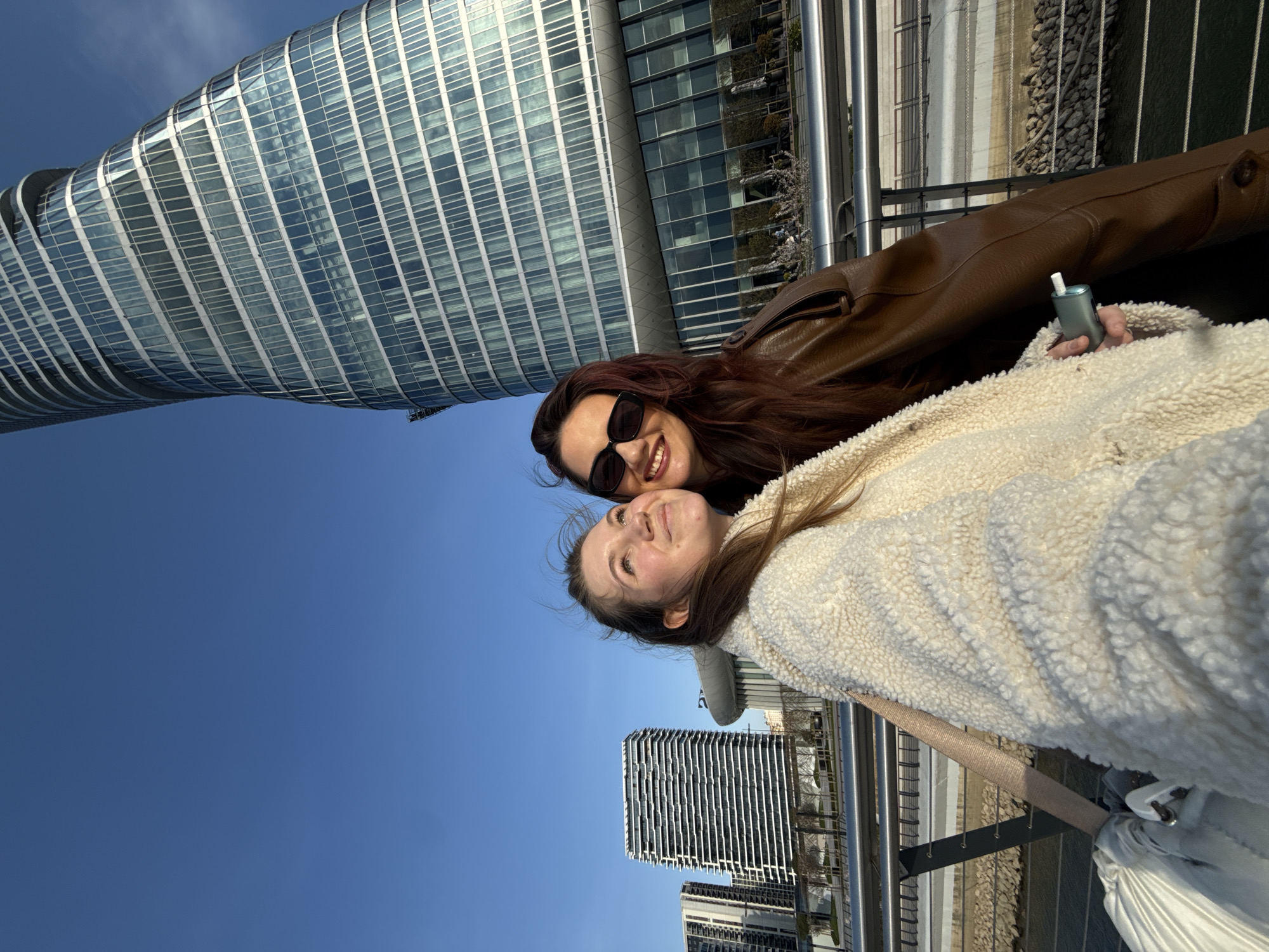 Two females are captured in a close-up, outdoor photograph in Belgrade, Serbia, specifically within the Belgrade Waterfront development. The Kula Belgrade (Belgrade Tower), a prominent modern skyscraper with a twisting glass facade, is visible in the background on the right under a clear blue sky. Other contemporary multi-story buildings are seen further left in the background.
In the foreground, a female on the left with long light-brown hair, wearing a cream-colored sherpa jacket, gazes upwards and to her right with a slight smile. She holds a light blue electronic device with a white tip in her right hand, positioned between herself and the second female. The second female, on the right, has long dark reddish-brown hair, wears dark sunglasses and a brown leather jacket, and smiles directly at the camera. A metallic railing runs horizontally behind the subjects, separating them from the background which includes a body of water (likely the Sava River) with rocky banks and landscaped areas. Sunlight illuminates the subjects from the front, indicating a sunny day.