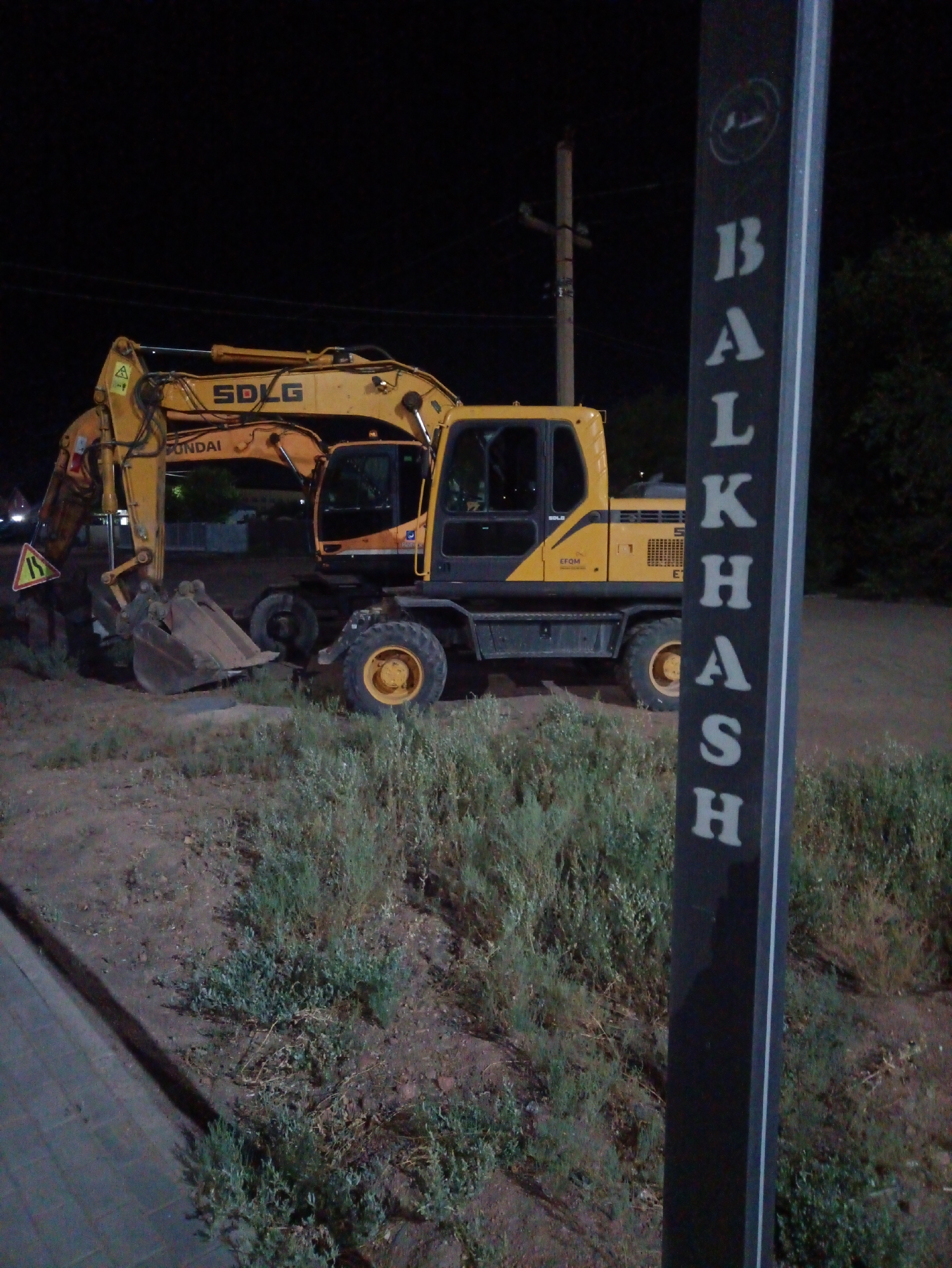 An excavator is parked on a dirt and grass area at night. The excavator is yellow and has "SDLG" and "HYUNDAI" logos visible. A vertical black sign with white lettering is in the foreground, to the right of the excavator, displaying the word "BALQAŞ" vertically. The sign also contains a circular emblem at the top. In the background, a utility pole and some trees are visible against a dark sky. A warning sign with a road symbol is attached to the excavator's arm. The ground appears uneven, with some patchy vegetation.