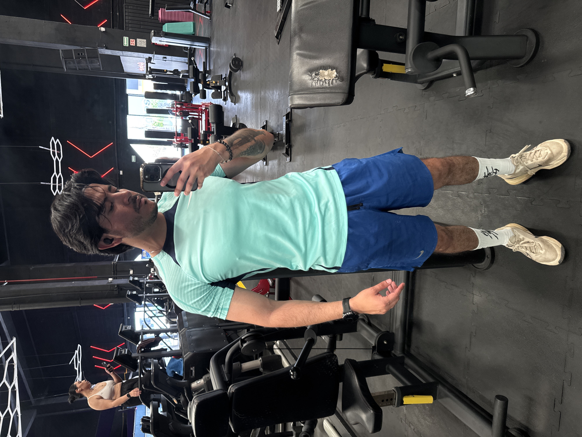 A man is taking a selfie in a gym. He is wearing a light blue t-shirt, blue shorts, white socks, and white athletic shoes. He is holding a smartphone in his left hand. He has dark hair and a beard. Behind him is gym equipment, and in the background, there is another person exercising. The gym appears to be located in Puebla, Mexico, based on the location information.