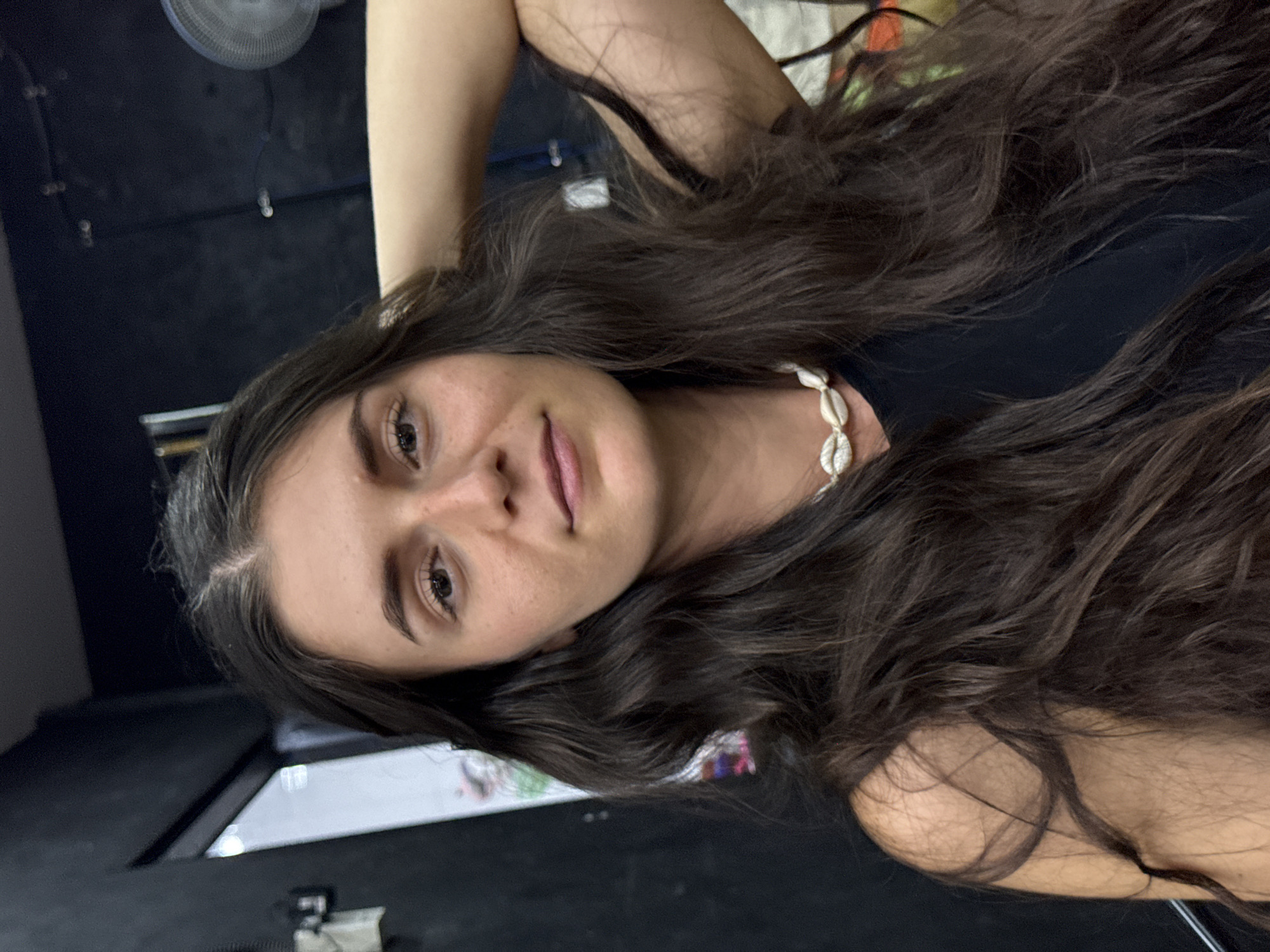 A close-up portrait, originally rotated 90 degrees clockwise, features a young woman with long, dark, wavy hair, looking directly at the camera. Her head is tilted slightly, and her right arm is raised, with her hand likely behind her head or neck, causing her abundant hair to cascade around her shoulders and chest. She has light brown eyes, defined eyebrows, and appears to be wearing a light pink lip product. Her expression is neutral with a soft, subtle hint of a smile or smirk.
She is wearing a simple black top, possibly a tank top or camisole. A distinctive necklace made of white cowrie shells is visible around her neck, adding a natural, bohemian touch to her attire. Her skin appears clear with a few small freckles or moles visible on her face.
The background is predominantly dark and blurred, suggesting an indoor setting. In the upper left corner, a small, circular electric fan is partially visible against a dark wall or ceiling. Towards the lower left, some blurred reflections or light sources suggest the presence of other objects or surfaces in the room, possibly a screen or table. The overall lighting on her face is soft and direct.
The scene depicts a casual portrait moment in an indoor environment, consistent with a relaxed setting in Phu Thuy Ward, Vietnam. The presence of the fan could suggest a warm climate. No visible text is present in the image.