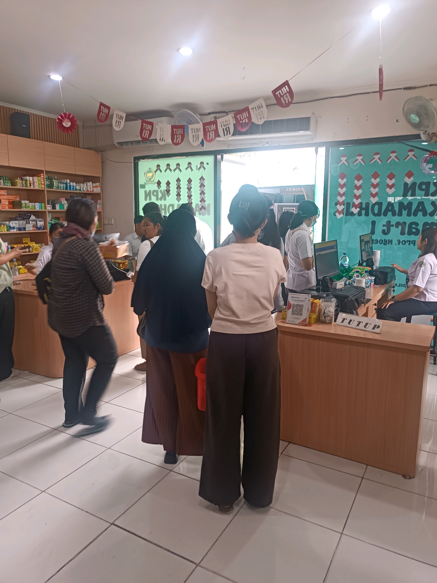 The image depicts the interior of what appears to be a pharmacy or a retail store, possibly located in Denpasar, Indonesia, given the context. The scene is bustling with activity, with several individuals present. In the foreground, a person is seen from behind, wearing dark pants and a light-colored t-shirt. To the left, another person in a plaid shirt and dark pants is walking away, holding a box. Further into the store, there's a counter where several people are gathered, some appearing to be staff and others customers. One staff member in a white uniform is interacting with a customer. The background shows shelves stocked with various products, suggesting it's a retail environment. Decorative banners with Indonesian text, possibly related to a celebration or event, are hung from the ceiling. The lighting suggests an indoor setting, and the floor is tiled.