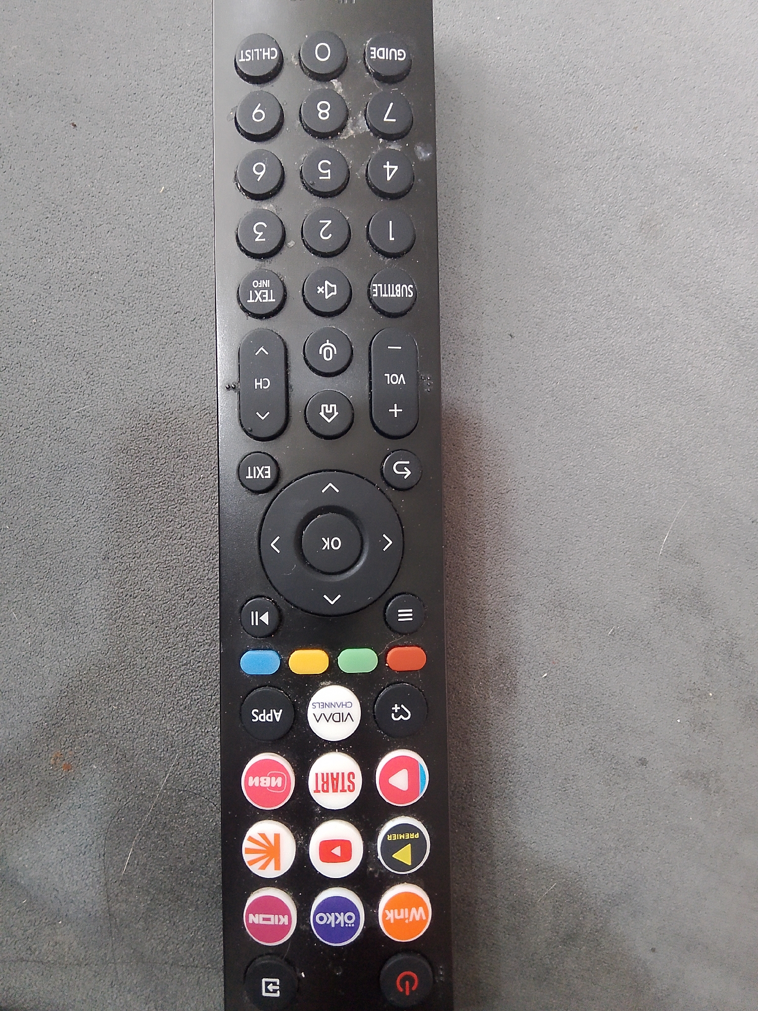 This is a close-up, overhead photograph of a black television or smart device remote control, positioned vertically on a speckled, dark grey surface. The remote is visibly used, with dust accumulated on its buttons and casing.
The remote features a comprehensive array of buttons, including:
- A numeric keypad from 0 to 9.
- Standard control buttons such as CH.LIST, GUIDE, TEXT INFO, SUBTITLE, VOL (+ and -), CH (up and down arrows), EXIT, and a central circular OK button surrounded by directional arrows.
- Media playback controls, including a button for PLAY/PAUSE (represented by two vertical lines and a play triangle).
- Additional functional buttons include APPS, a mute symbol, a back/undo arrow, and a menu symbol (three horizontal lines). Four distinct colored rectangular buttons (blue, yellow, green, and red/orange) are also present.
A prominent section of the remote is dedicated to direct access buttons for various streaming services and platforms, indicated by their logos and text:
- VIDAA CHANNELS
- ИВИ (IVI)
- START
- KION
- OKKO
- Wink
- PREMIER (represented by a yellow triangle on a black button).
- There is also a red circular button with a white play symbol, a white circular button with the YouTube logo, and another white circular button featuring a stylized orange logo.
The power button, marked with the universal power symbol, is located at the bottom right of the remote. The background is an unadorned, textured grey surface, providing no additional context about the location or environment.