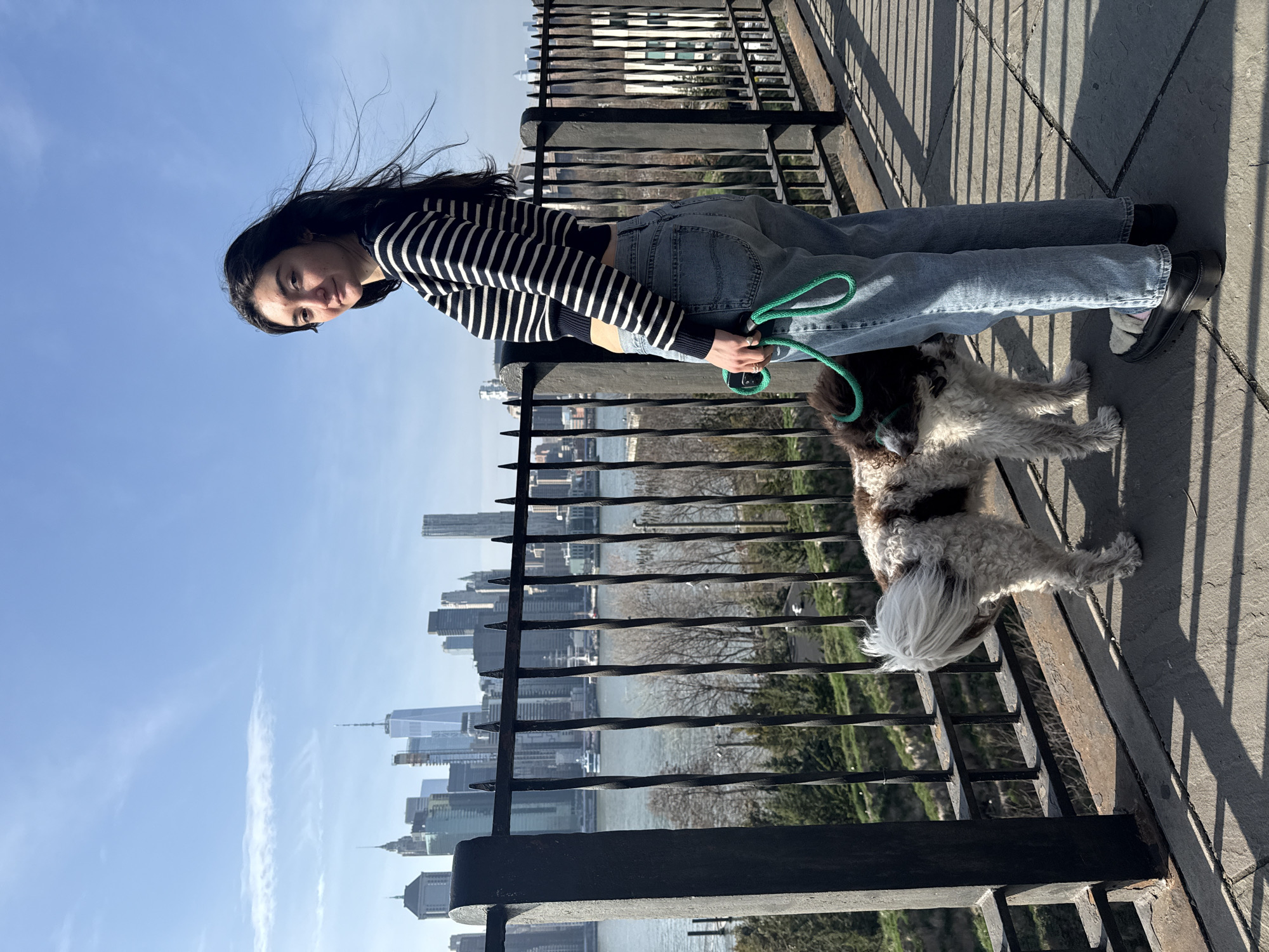 A young woman wearing a black and white striped shirt and light blue jeans holds a green leash connected to a medium-sized dog with brown and white fur. They are standing on a concrete platform behind a black metal railing. Behind the railing is a view of the New York City skyline, including the One World Trade Center building. The day is sunny with a clear blue sky. The location appears to be a park or promenade overlooking the city.