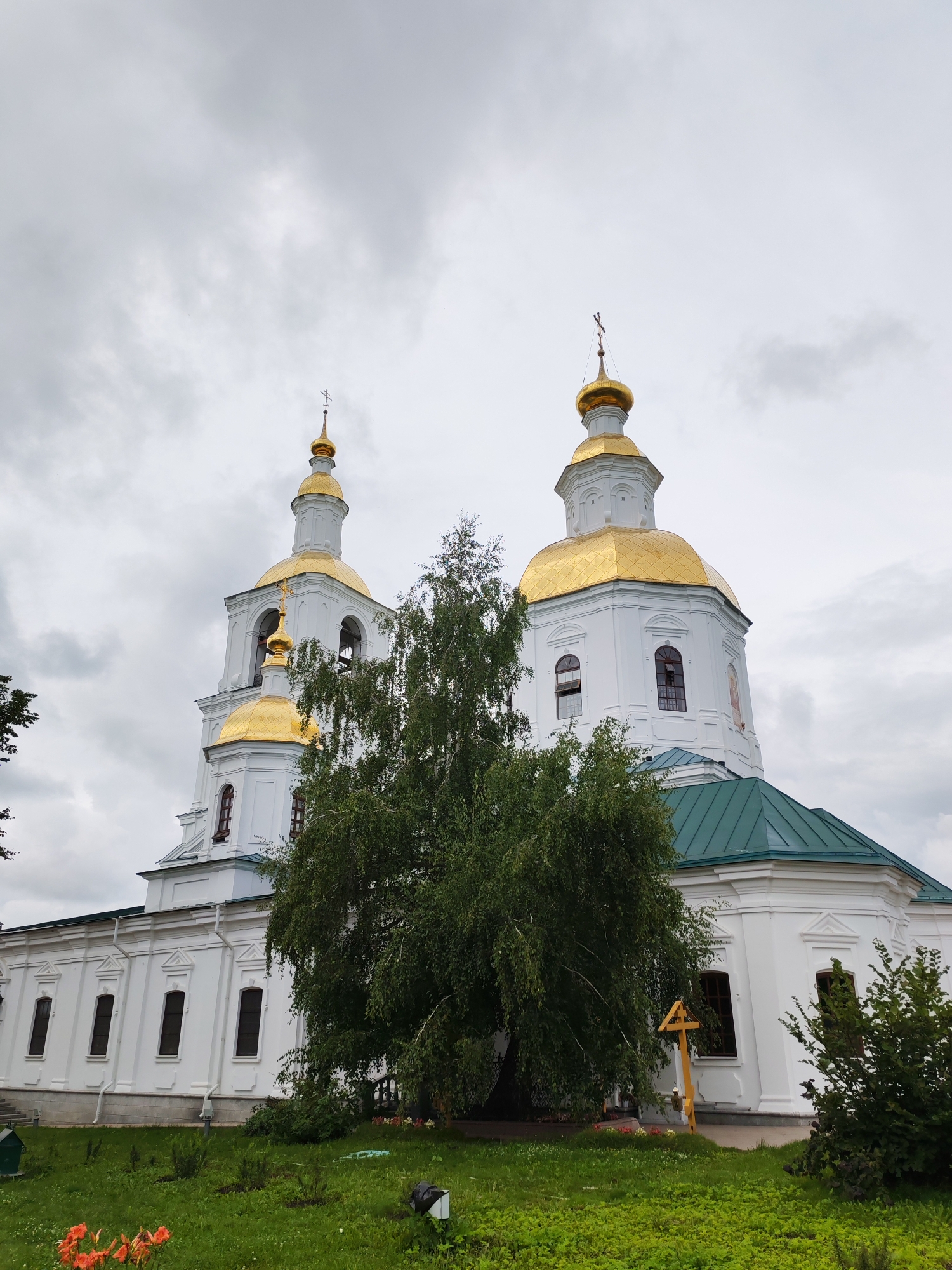 The image shows a white church with golden domes, located in the city of Diveyevo, Russia. The church has two bell towers and a main dome, all topped with crosses. A lush green tree stands in front of the church, partially obscuring the view of the bell towers. The church is surrounded by a well-maintained lawn and flower beds, with a wooden cross visible on the right side of the image. The sky is overcast with gray clouds.