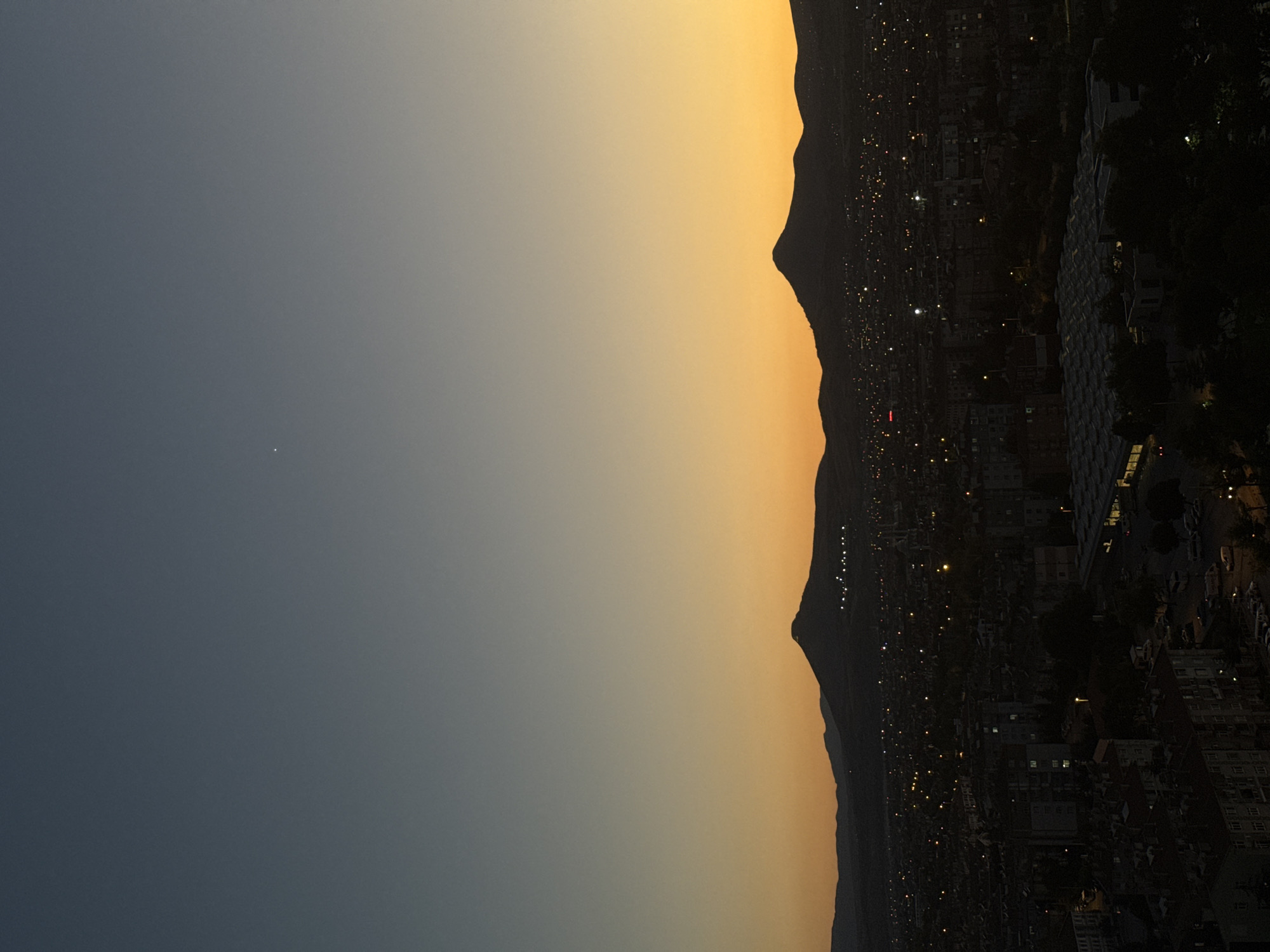 This photograph captures a panoramic view of Konya, Turkey, at dusk. The sky transitions from a deep twilight blue at the top to a vibrant orange and yellow gradient near the horizon, suggesting the last light of the setting sun. Silhouetted against this colorful sky are two mountain ranges. The foreground is dominated by the cityscape of Konya, with numerous buildings, streets, and vehicles visible. Streetlights and illuminated windows dot the urban landscape, creating a warm glow against the darkening buildings. A large, modern structure with a distinctive patterned roof is prominent in the lower right portion of the image. The overall scene is one of a city settling in for the evening, with the natural beauty of the twilight sky providing a dramatic backdrop. There are no people or specific actions clearly depicted, rendering the image a static observation of the urban environment at a specific time of day.