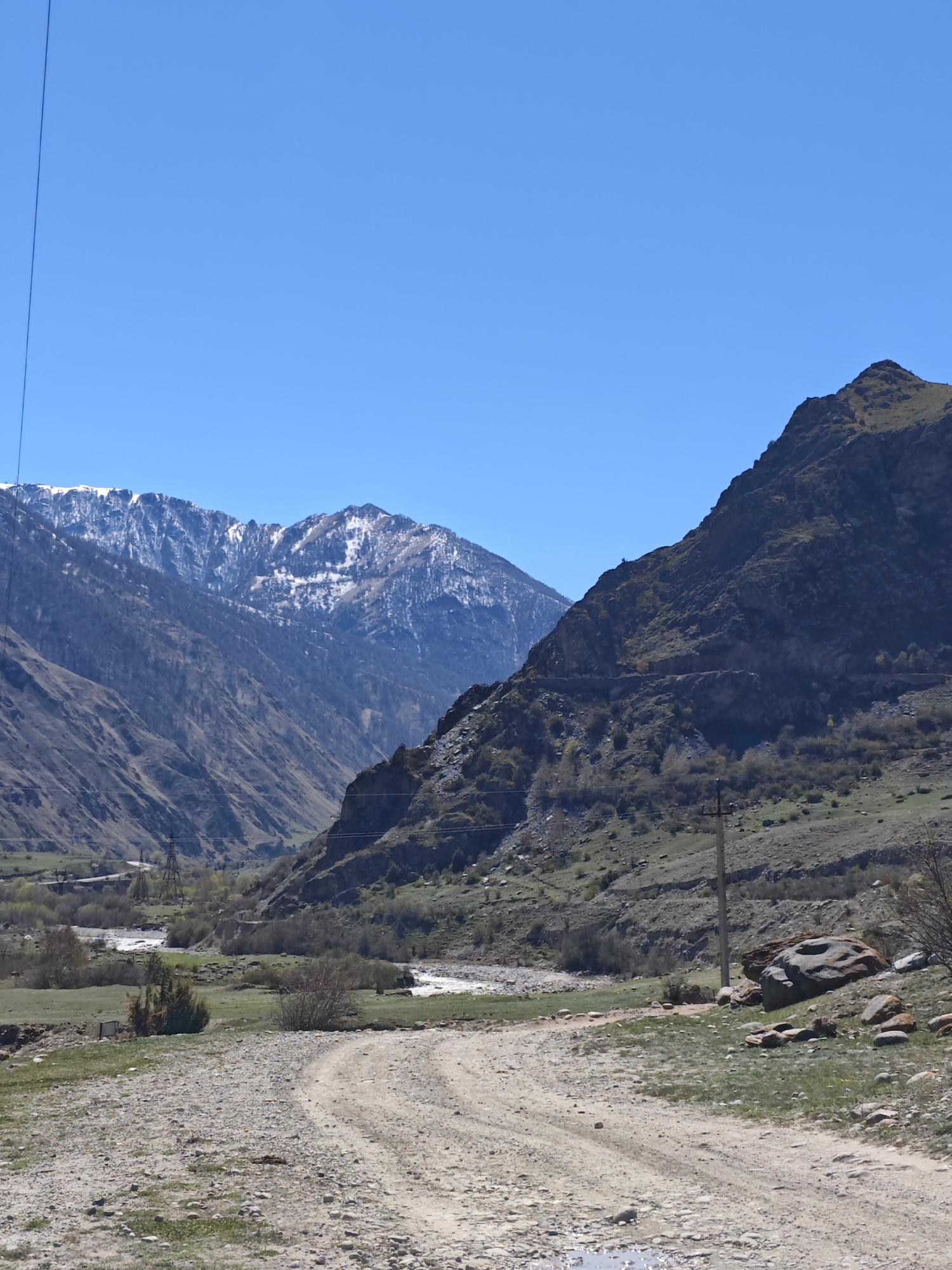 The image shows a dirt road leading into a valley in Bylym, Russia. Snow-capped mountains are visible in the distance. A river runs through the valley, with power lines and electrical towers present on either side of the valley. Boulders are present along the right side of the road. The sky is clear and blue. The vegetation consists mostly of grass and sparse trees.
