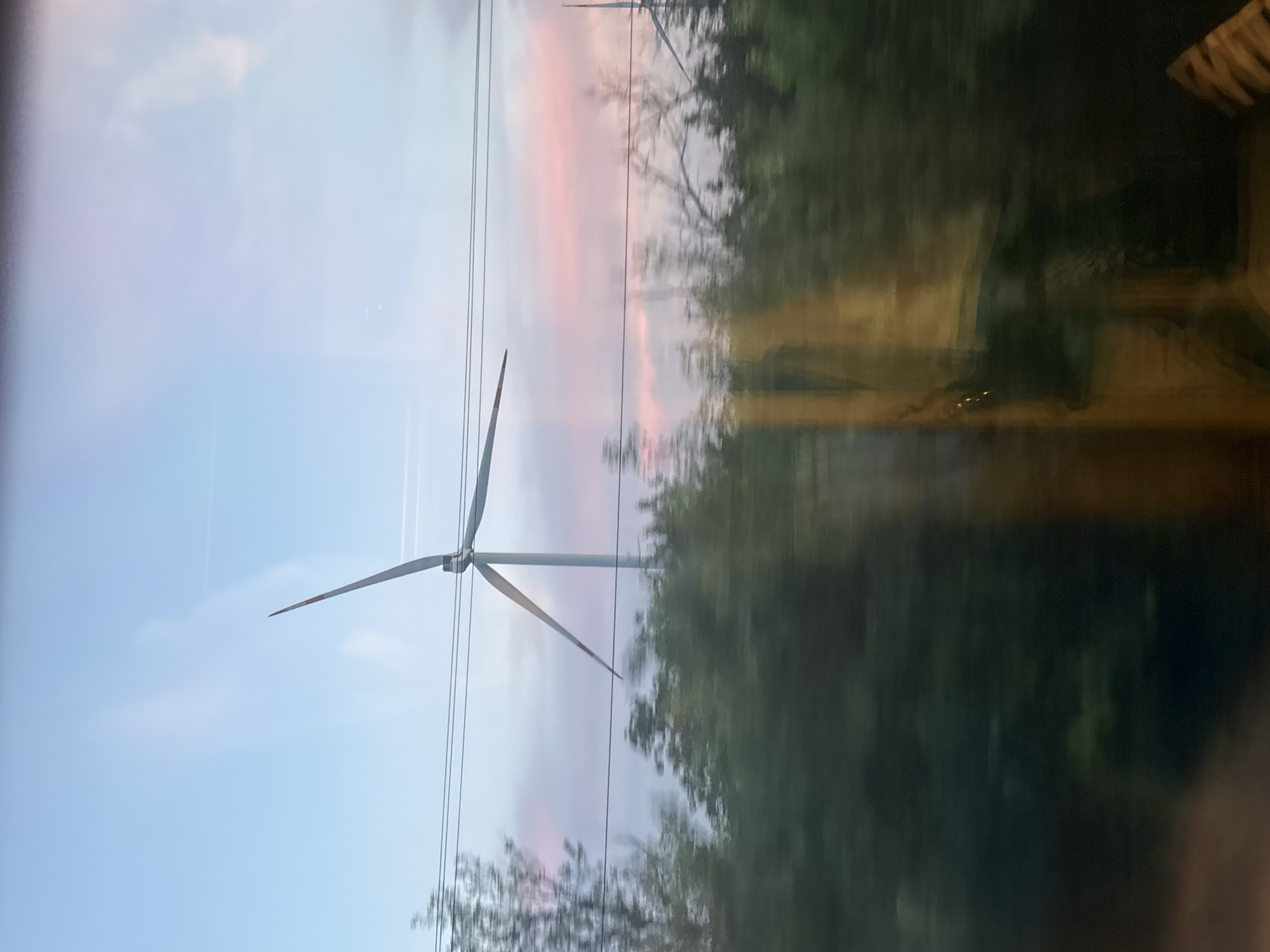 The image captures a wind turbine against a sky with hues of blue and pink, suggesting either sunrise or sunset. The turbine's blades are angled, with one clearly visible extending upwards and to the right, and another partially visible extending to the left. Power lines stretch horizontally across the frame, passing in front of the turbine. Below the power lines, a dense line of trees forms a dark green border, with some of the foliage appearing blurred due to motion. The foreground is heavily obscured by reflections on a window, displaying warm yellow and brown tones, and the dark, blurred shapes of what appear to be interior elements. The overall scene suggests a landscape view observed from a moving vehicle. There are no people or animals visible. The location of Verkhnegoveinyi, Russia is not directly evident from the visual information in the image.