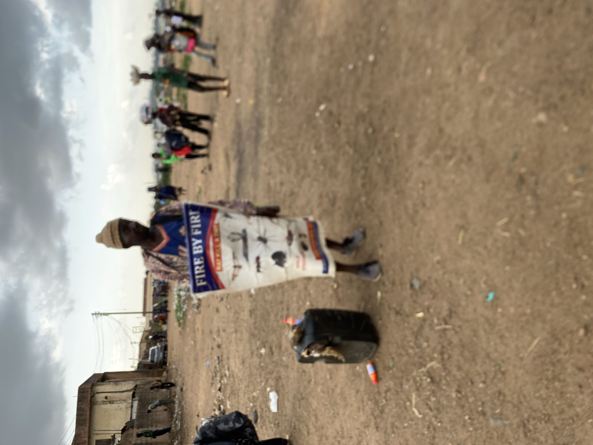 A person stands in a sandy, trash-strewn area in Bukuru, Nigeria, wearing a hat, a patterned shirt, and carrying a large, white, rectangular bag that reads "FIRE BY FIR" on its side. In front of the person, on the ground, is a damaged black container with debris spilling out. Scattered around are various pieces of trash, including plastic bottles and bags. In the background, several other people can be seen further down the sandy area. Behind the person, a building is visible, partially obscured by trash and debris. Overhead, a cloudy sky dominates the view.