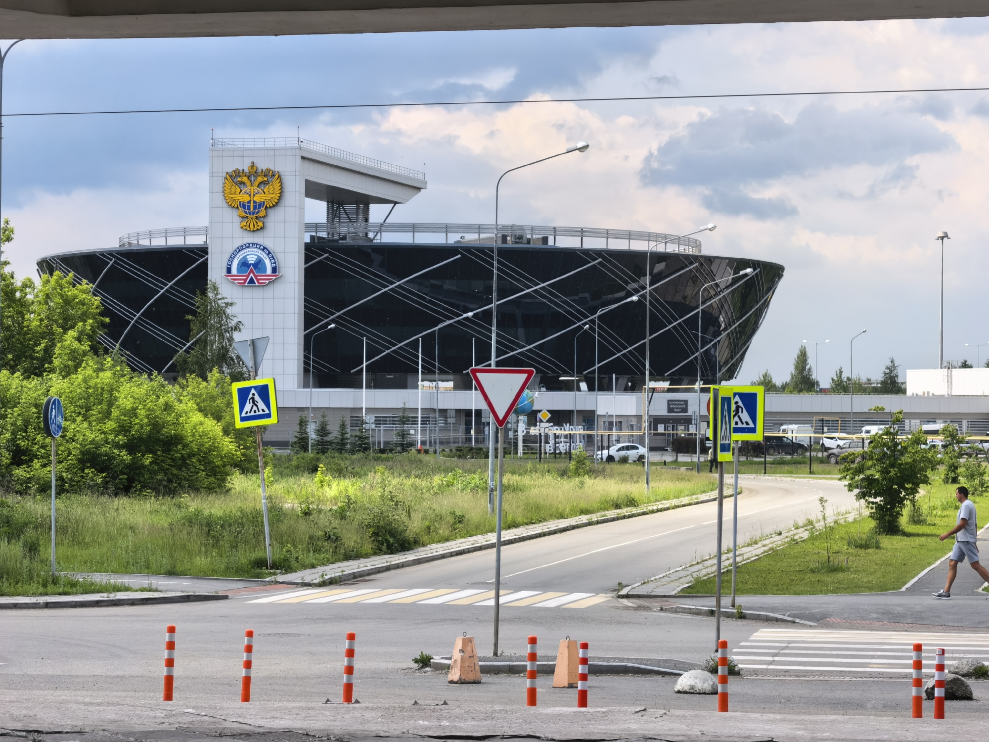 In an outdoor, daytime scene, a modern, futuristic-looking building with a black, curved facade dominates the background, topped with a white structure featuring a large golden double-headed eagle emblem and a circular logo below it. The building appears to be an official or governmental institution. In the foreground, a paved road curves to the right, marked with a pedestrian crossing. Orange and white striped bollards are strategically placed along the road. Several traffic signs are visible, including a blue circular sign with a bicycle icon, a yellow square sign with a pedestrian symbol, and a red and white inverted triangle sign. Green grass and trees surround the road and building. A man in a light grey t-shirt and shorts walks across the pedestrian crossing from right to left. The sky is partly cloudy.