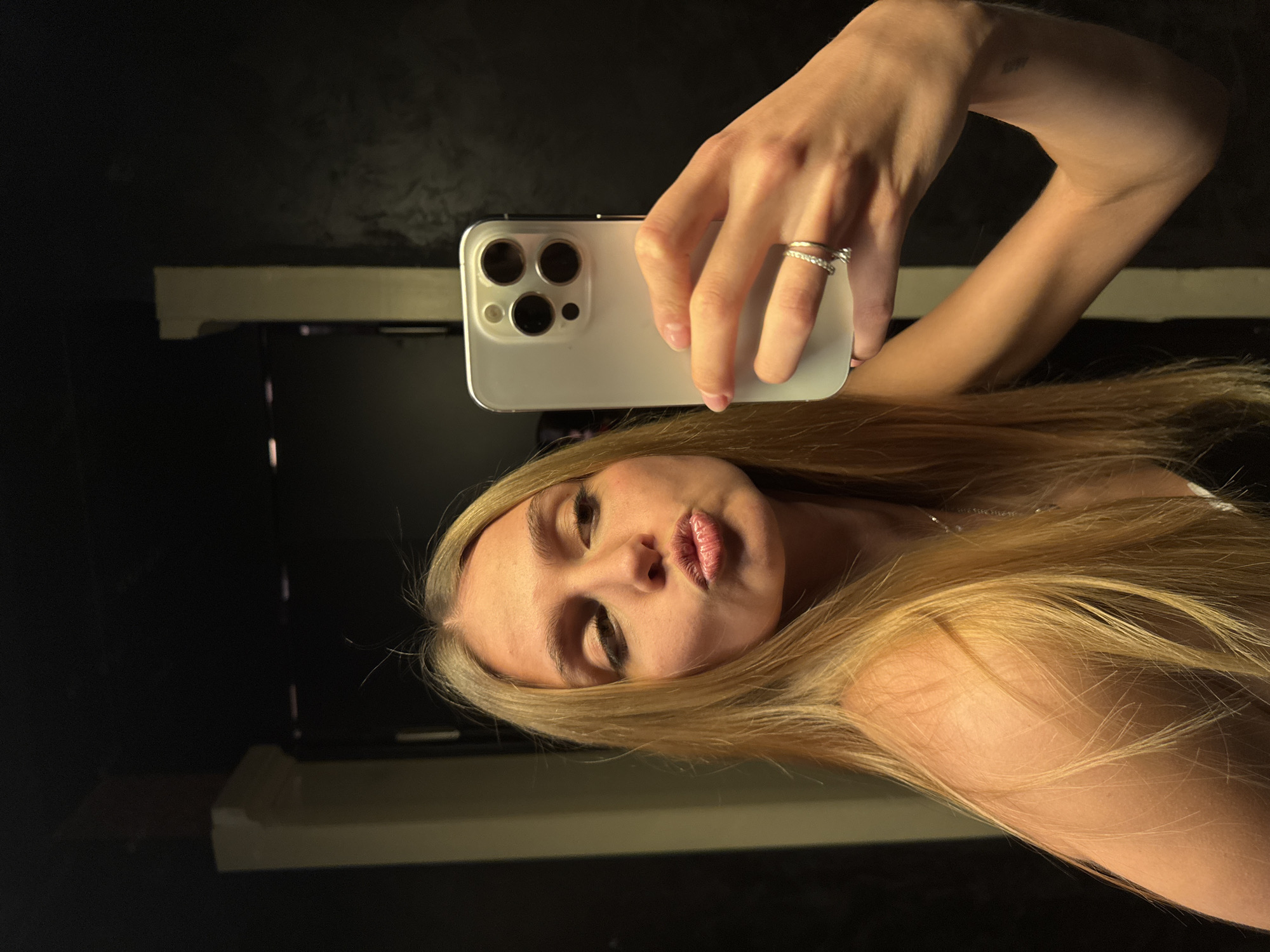 A selfie image captures a young woman with blonde hair taking a picture of herself in what appears to be a dimly lit room, possibly a bathroom. She is holding a white smartphone with multiple camera lenses up and to her right, slightly tilted. Her hair is flowing across her shoulder and arm. She is wearing a ring on her ring finger. The background consists of dark walls and a reflective surface, possibly a mirror, showing blurred shapes of what could be a counter or sink. The location is unknown, but the request includes that this image was taken in the city of Tbilisi, Georgia.