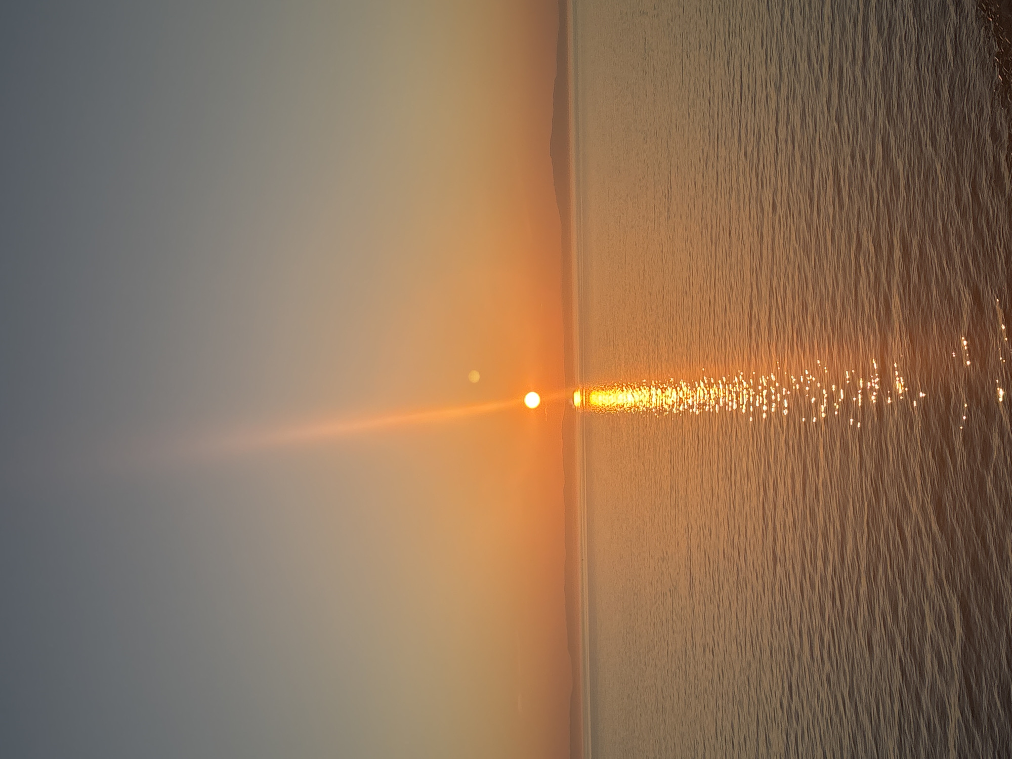 The image depicts a tranquil sunset over a body of water. The sun, a brilliant orb of orange, is positioned near the horizon, casting a radiant glow across the scene. A luminous reflection of the sun shimmers on the water's surface, creating a path of light. In the distance, a silhouette of hills or mountains can be seen against the warm hues of the sky. The sky itself transitions from a soft blue at the top to a warm orange near the horizon, a typical characteristic of a sunset. The water appears calm, with gentle ripples disturbing its surface. The foreground shows a dark, rocky embankment. This scene is located in the city of Turka, Russia.