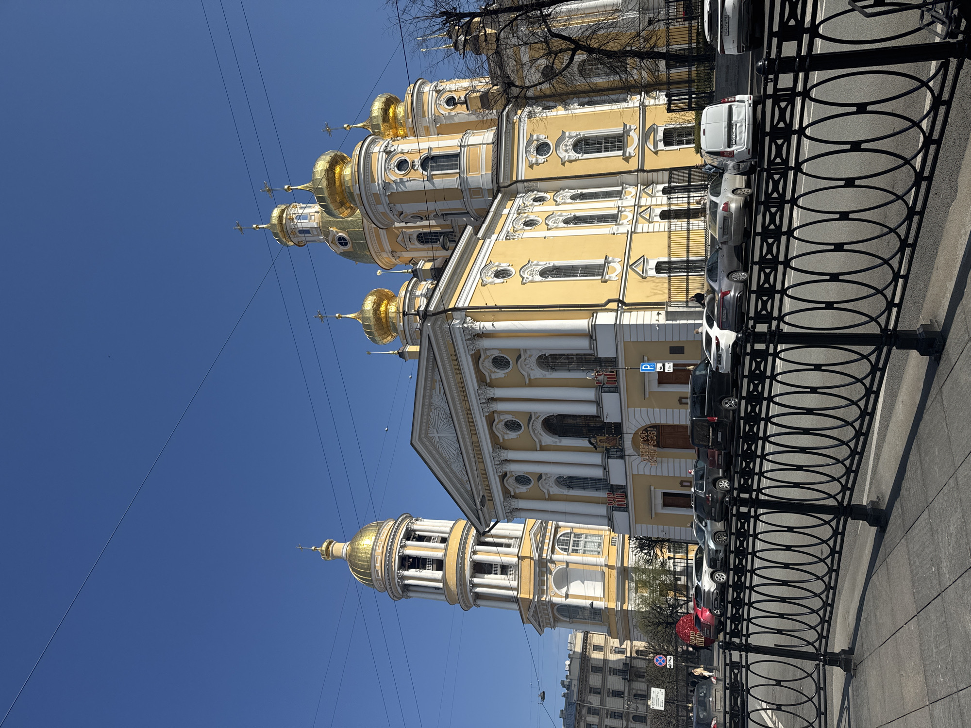 The image depicts the Catherine Church on Vasilyevsky Island in Saint Petersburg, Russia. The church is a yellow and white baroque building with golden domes, visible against a clear blue sky. A black metal fence with circular and cross-shaped designs runs along the sidewalk in the foreground. Several cars are parked along the street next to the church. Bare tree branches are visible in the upper right corner of the image. Pedestrians and other buildings are visible in the distance.