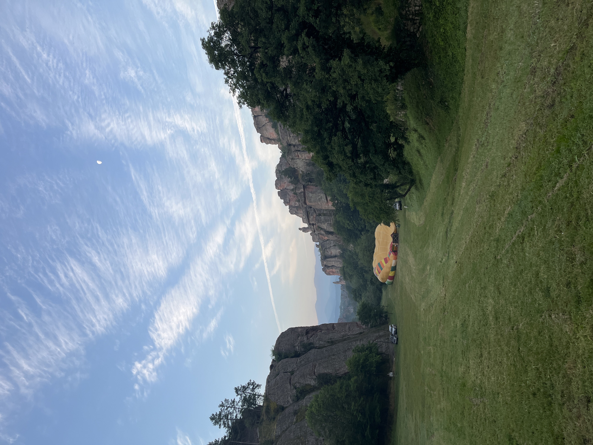 This wide-angle outdoor image captures a picturesque valley or open field nestled between imposing rock formations under a soft, clear sky. The ground is covered in vibrant green grass, exhibiting streaks from recent mowing.
The sky, a serene light blue, is adorned with numerous wispy, white cirrus clouds that stretch diagonally across the frame. A distinct crescent moon is visible in the upper right quadrant, alongside a faint, linear contrail from an aircraft crossing horizontally in the mid-sky.
On the left side of the valley, a towering, steep, grey-brown rock cliff rises dramatically, featuring a few dark green pine trees along its upper edge. At the base of this cliff, two light-colored utility vehicles, possibly SUVs or vans, are parked on a dirt path.
In the mid-ground, a large hot air balloon is laid out on the grassy field. Its envelope is predominantly yellow, detailed with prominent red and green horizontal stripes near its base, indicating it is either in the process of inflation or deflation.
The right side of the valley is characterized by additional large, weathered rock formations, partially obscured by a dense canopy of dark green trees in the foreground. In the distant background, hazy blue mountains or hills are visible between the rock formations, suggesting an expansive landscape.
The overall lighting is soft and diffused, consistent with either early morning or late afternoon conditions. No discernible human figures are visible in the scene, and the location is identified as the city of Unknown, Unknown.