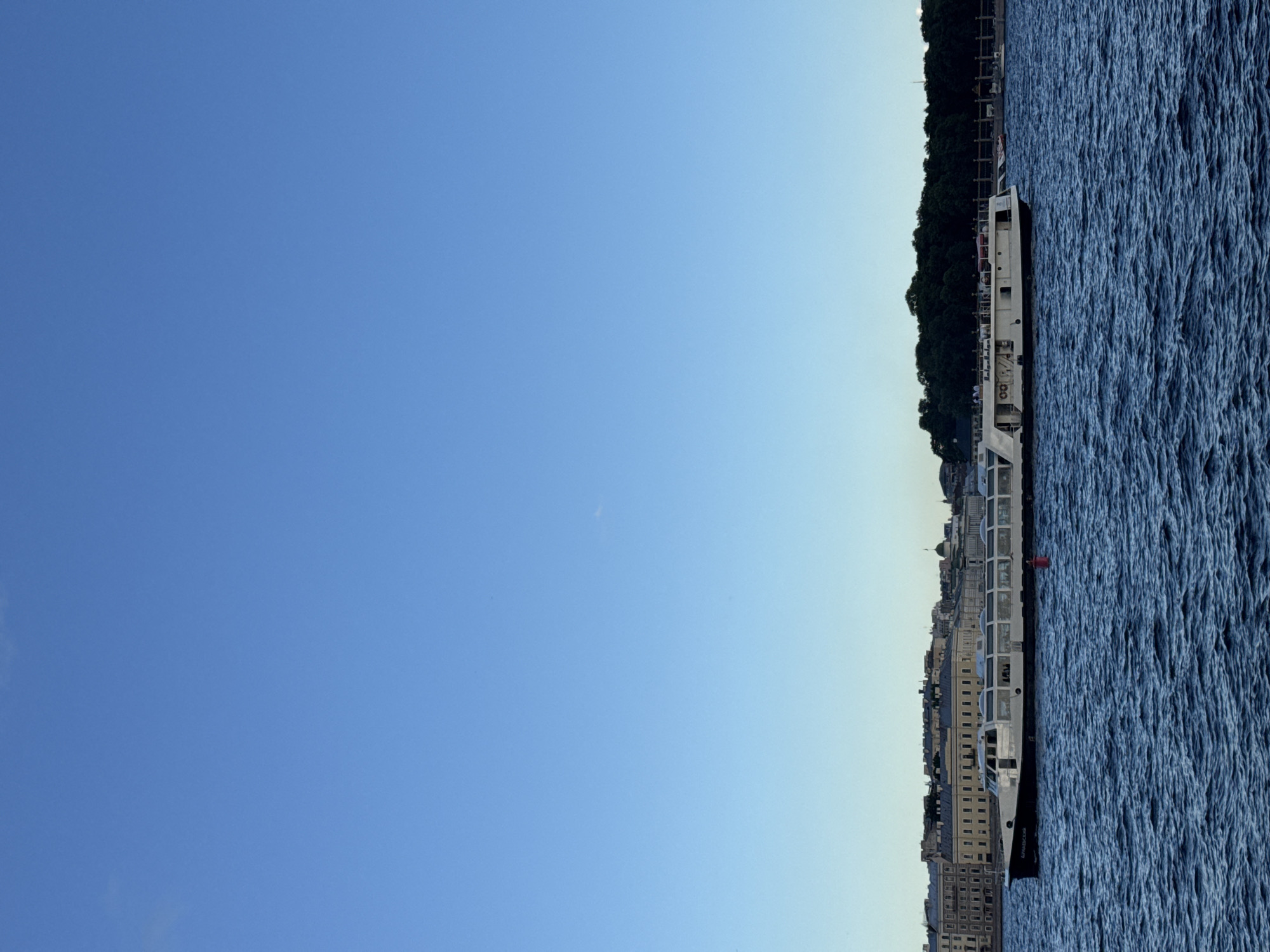 The image captures a scenic view of the Neva River in Saint Petersburg, Russia, under a clear blue sky. A long, white passenger boat with a black hull navigates the choppy, dark blue water. The boat features numerous windows, suggesting it is designed for sightseeing tours, and people can be seen on its upper deck. In the background, a line of historic buildings with ornate facades and a domed structure are visible along the riverbank, hinting at the city's rich architectural heritage. Lush green trees form a dense backdrop to the buildings. A red buoy floats in the water near the boat. The overall scene conveys a sense of calm and timeless beauty, characteristic of Saint Petersburg's waterfront.