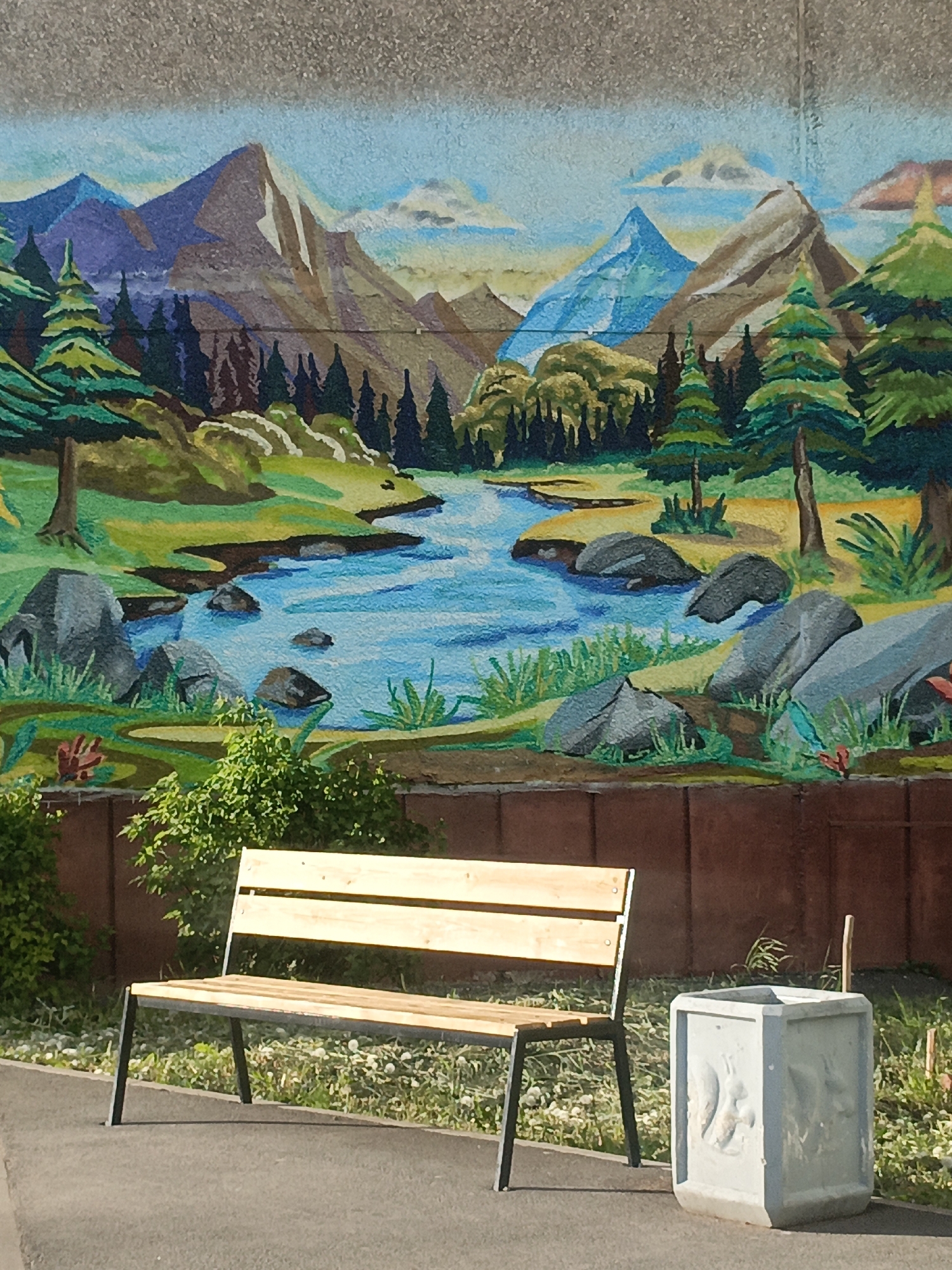 The image shows a bench and a concrete planter in front of a mural depicting a mountain landscape with a river. The mural is painted on a concrete wall. The bench is made of wood and metal, with a light-colored wooden seat and a dark metal frame. The concrete planter is square and has some markings or designs on its surface. The scene appears to be outdoors, possibly in a park or public area. The photo may have been taken in Ufa, Russia.