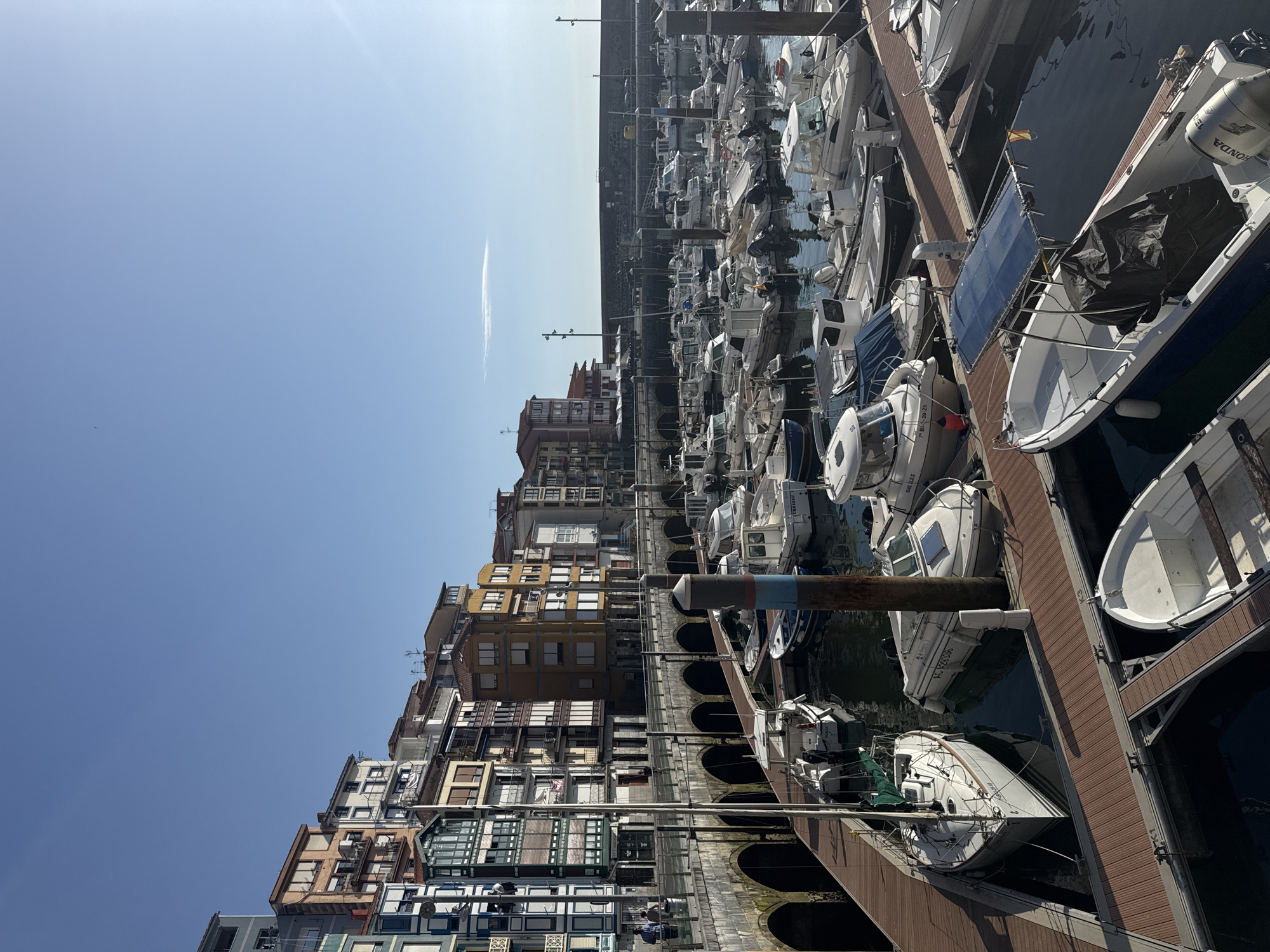 This image captures a scenic view of the port of Bermeo, Spain, under a clear blue sky. The foreground features a wooden dock with several boats moored. Among them is a white sailboat with a green sail, and various motorboats of different sizes, some with protective covers. The middle ground showcases more boats in the marina, reflecting in the calm water. In the background, a series of colorful buildings, characteristic of coastal Spanish architecture, line the waterfront. These buildings, with their varied facades and balconies, are perched above a stone arcade. A few people are visible walking along the promenade in front of the buildings. The scene is bathed in sunlight, creating a peaceful and picturesque maritime atmosphere.