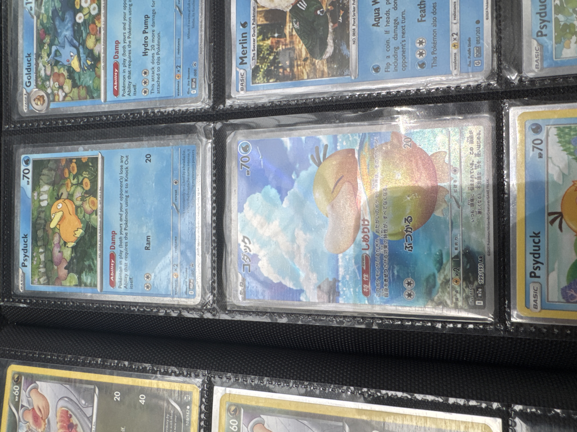 The image shows a close-up, eye-level view of a black binder filled with Pokemon cards. The binder is oriented vertically. Several Pokemon cards are visible within the plastic sleeves of the binder. On the top row, from left to right, are parts of three cards: the first is largely obscured but shows the top of a yellow border and the number "HP 60", the second card features a bright blue background with an orange Pokemon character and the text "Psyduck" and "70 HP", and the third card has a blue background, an orange character, and the text "Golduck" and "70 HP". The middle row displays two cards. The card on the left has a predominantly blue sky and ocean background with a character that appears to be a light orange duck-like Pokemon. The text on this card is in Japanese. The card on the right features a light blue background with a character that looks like a green duck wearing a tracksuit. The text on this card includes "Merlin" and "The Soccer Duck Pokémon". The bottom row shows at least one card with a yellow border and the text "Psyduck" and "70 HP". The lighting suggests it is indoors, possibly a home or a store. The image does not provide any information about the city of El Paso, Texas, or the United States of America.