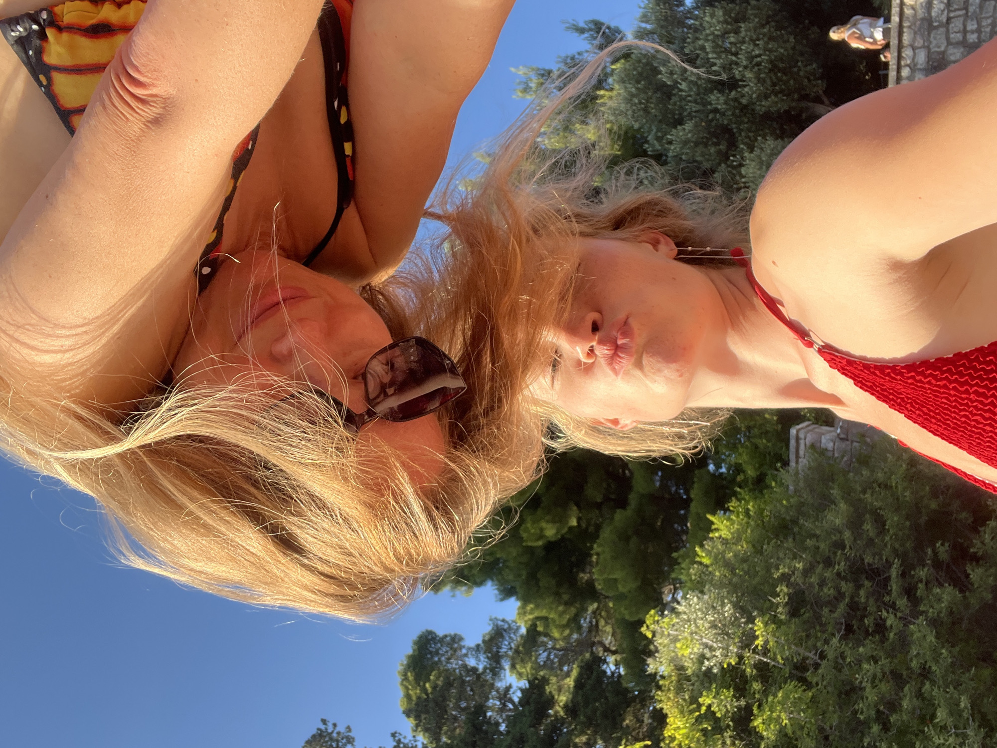 This image depicts two individuals, appearing to be women, engaged in a playful interaction outdoors in Rađenovići, Montenegro. The scene is illuminated by strong natural sunlight under a clear blue sky, surrounded by dense green foliage, predominantly trees.
In the upper portion of the frame, one individual is inverted, bending forward with long, blonde hair flowing downwards and partially obscuring her face. She wears a bikini top featuring a prominent orange and black butterfly wing pattern. Dark-framed sunglasses are perched on her head, visible near her right ear. Her visible features suggest a playful or smiling expression.
Below and slightly to the right, a second individual stands upright, looking upwards towards the first. This individual has blonde hair, with some strands blowing across her face. She wears a red, textured bikini top and long, thin, red dangle earrings. Her lips are pursed in a distinct pouting or "kissing" expression.
In the far background, behind the upright individual, a stone wall or structure is visible, with a blurry figure of another person seated or standing upon it. The overall environment suggests a natural, warm-weather setting with ample vegetation.