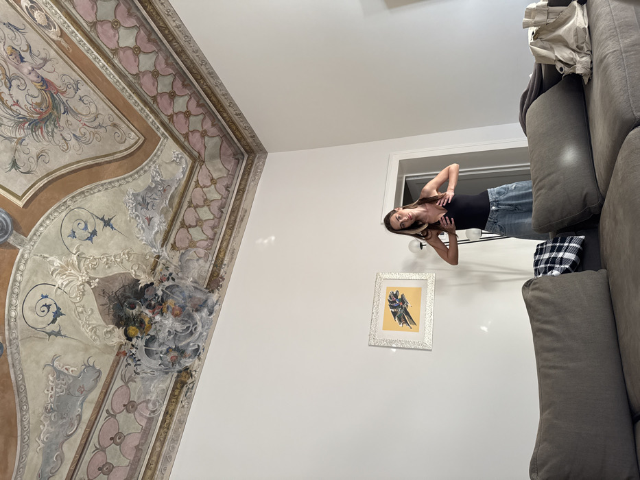 The image depicts an indoor scene within a room in Venice, Italy, characterized by a high, ornately frescoed ceiling and plain white walls. The ceiling features a central painted area with swirling patterns, light brown borders, and a repeating frieze pattern of interlocking pink and light green shapes. A prominent detail on the ceiling is a painted and possibly stuccoed vase overflowing with colorful flowers and foliage.
In the mid-ground, a female individual stands facing the camera. She has long, straight dark brown hair with lighter streaks. She is wearing a black sleeveless v-neck top and light-wash, loose-fitting denim jeans. Her hands are positioned on her chest, appearing to adjust her garment. Her expression is neutral.
To the left of the individual, on the white wall, a framed artwork hangs, featuring a dark, possibly abstract or stylized, shape against a yellow background, enclosed in a silver-colored textured frame. A tall, white-globed floor lamp is partially visible behind the woman. A plain white doorway is visible beyond the woman, leading into an adjacent room.
In the foreground, a large, dark grey sectional sofa occupies the lower portion of the frame. A black and white plaid blanket is draped over one armrest of the sofa. A light-colored bag rests on the far end of the sofa, to the right.