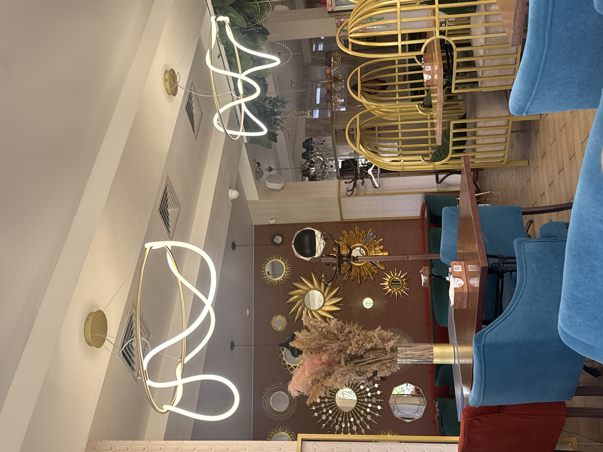 The image shows the interior of a restaurant in Chaikovsky, Russia. The scene includes tables and chairs, with blue velvet chairs positioned around a wooden table. Above, there are distinctive gold cage-like booths. Decorative lighting fixtures with circular, intertwining white light elements hang from the ceiling. The wall features a brown backdrop adorned with numerous decorative sunburst mirrors of varying sizes. A floral arrangement in a clear vase sits on a table. The interior design is a blend of modern and eclectic styles.