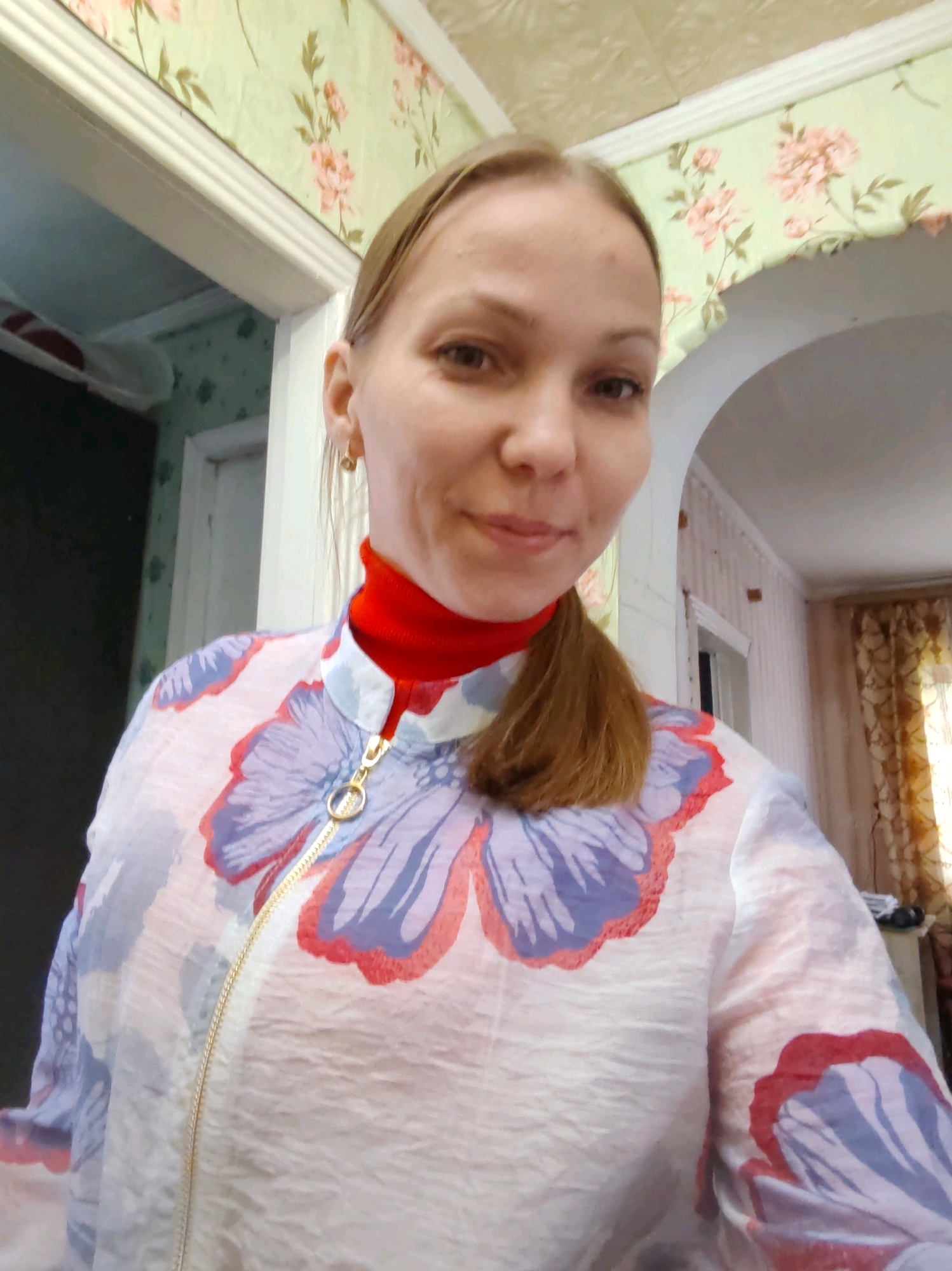 The image is a selfie of a woman in an indoor setting. She has fair skin, brown hair pulled back into a ponytail, and is wearing a red turtleneck and a white jacket with a floral pattern. The background shows floral wallpaper, a doorway, and curtains. The context suggests she is in a residential interior, possibly in the city of Chuhlom, Russia, given the information provided.
