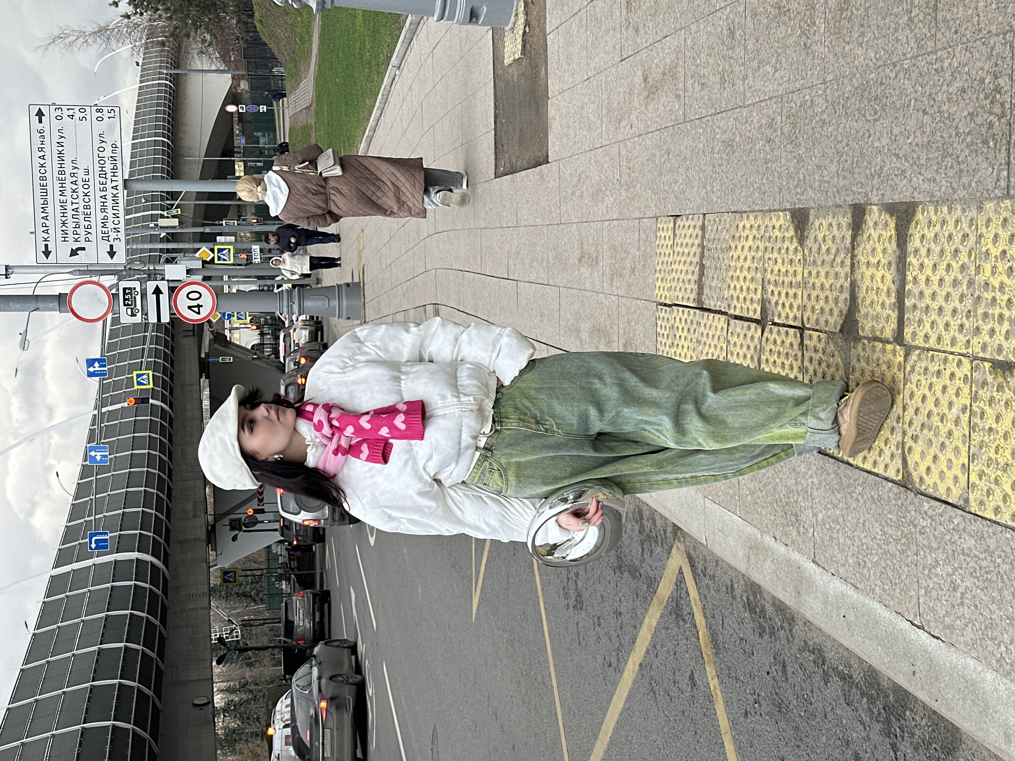 The photo shows a young woman in Moscow, Russia, standing on a sidewalk near a road. She is wearing a white jacket, green jeans, a white beret, and a pink scarf with hearts. She is holding a silver, round bag.
In the background, there is road traffic, an elevated road structure, and a sign with street names in Russian. The sign indicates directions and distances to various locations, including КАРАМЫШЕВСКАЯ наб. (Karamyshevskaya Embankment), НИЖНИЕ МНЁВНИКИ ул. (Nizhnie Mnevniki Street), КРЫЛАТСКАЯ УЛ. (Krylatskaya Street), РУБЛЁВСКОЕ ш. (Rublevskoe Highway), and ДЕМЬЯНА БЕДНОГО ул. (Demyana Bednogo Street). Other signs include weight restrictions (2.5 t) and a speed limit (40 km/h).
Several pedestrians are walking on the sidewalk behind her. The scene appears to be taken during the day.