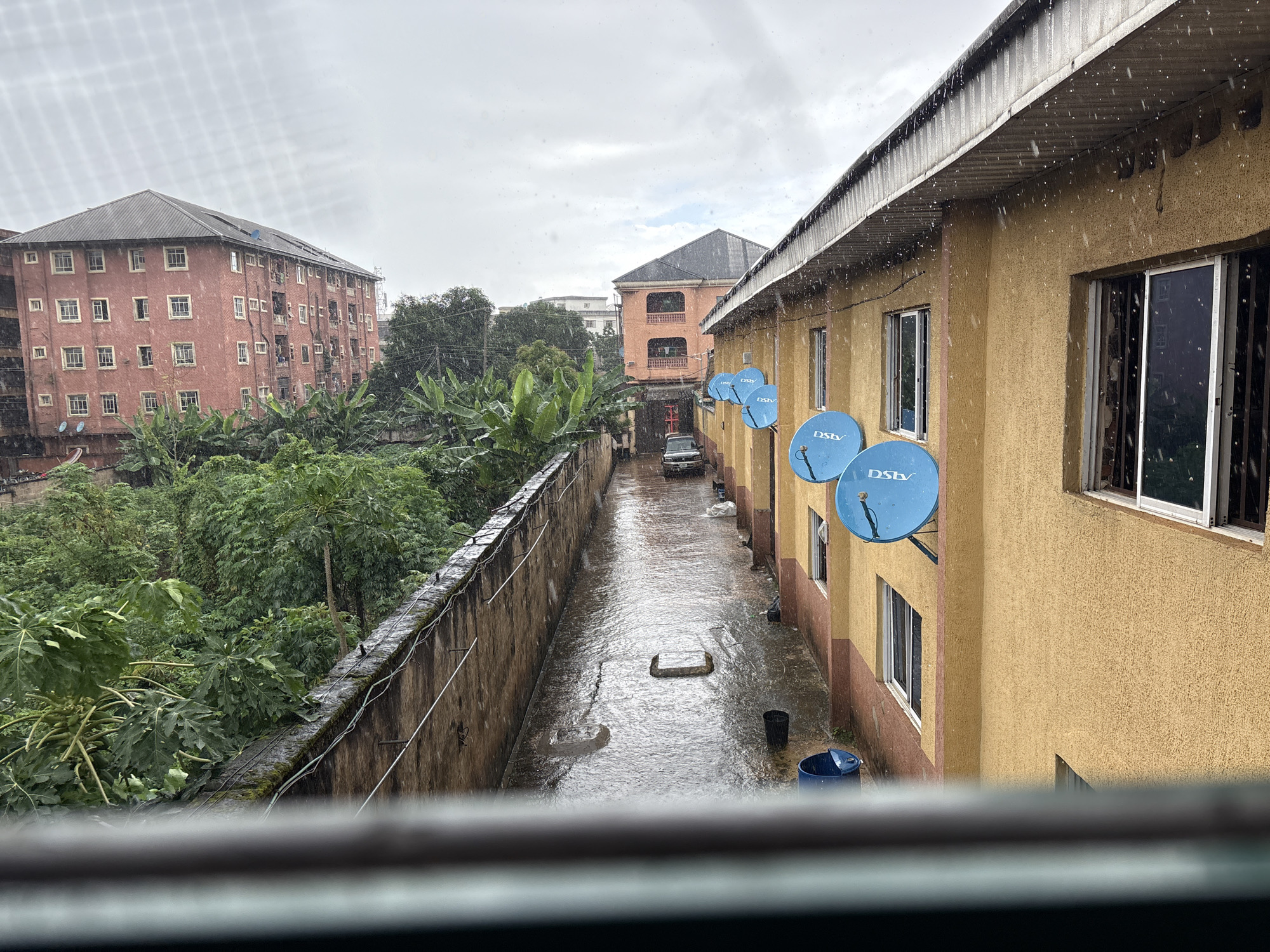 This media file depicts an outdoor scene in Onitsha, Nigeria, during what appears to be a heavy rain shower. The sky is overcast, suggesting either early morning or late afternoon, but the focus is on the wet conditions. The foreground is partially obscured by what seems to be a window frame.
The main subjects are the buildings and the environment. On the left, a tall, multi-story brick building with numerous windows stands against a backdrop of lush green trees and vegetation. In the center and right, a lighter-colored, two-story building with a corrugated metal roof is prominent. This building features several windows and is adorned with at least three blue satellite dishes, each marked with the "DSTV" logo. The path between the buildings is wet and reflects the sky, indicating standing water and active rainfall, with visible raindrops in the air and on surfaces.
The activity taking place is the rain itself, causing the ground to become saturated and the surfaces to glisten. There is a dark-colored car parked in the narrow walkway between the buildings. A blue plastic bin is visible near the wall of the second building, along with a black bucket. Two square concrete structures are also present in the wet walkway.
Notable details include the visible raindrops, the wet condition of the walkway, and the satellite dishes on the building, which are common sights in many urban Nigerian settings. The overall mood conveyed is one of a rainy day, likely bringing a sense of dampness and potentially interrupting outdoor activities. The location cues, such as the architecture of the buildings and the vegetation, along with the provided location context, firmly place the scene in Onitsha, Nigeria.