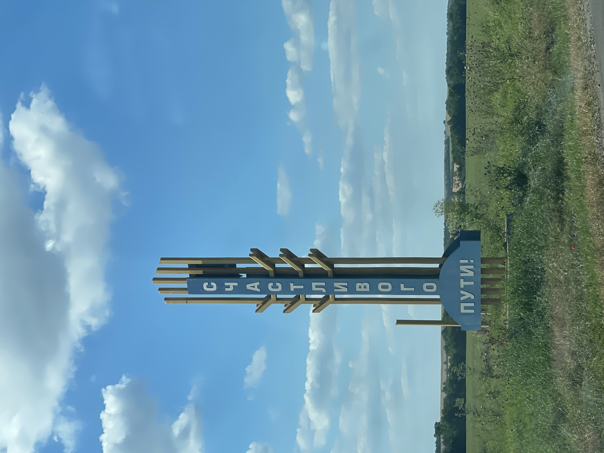 A vertical, blue and white sign with Cyrillic lettering stands in a grassy area under a partly cloudy blue sky. The sign is framed by vertical wooden posts and has a semicircular blue base. The top portion of the sign reads "СЧАСТЛИВОГО" vertically, and the base reads "ПУТИ!" horizontally. The surrounding environment consists of green grass, some wildflowers, and in the background, a treeline and fields under a bright, sunny sky with scattered white clouds. The location is identified as the city of Donskoe, Russia.