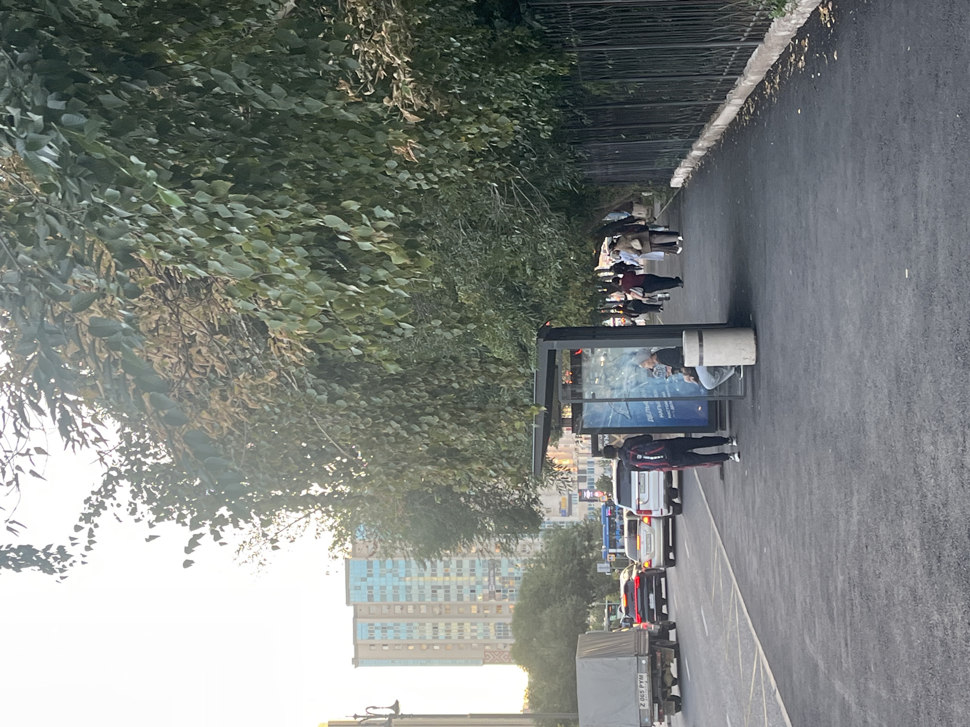 The image shows a street scene in Astana, Kazakhstan, during daylight. A tree-lined sidewalk runs alongside a road with several cars. On the sidewalk, a bus stop shelter is visible with a few people inside and waiting. A man with a backpack is walking away from the shelter. The road has a white line marking a lane divider and a yellow line marking the edge of the road. In the background, a tall building with blue-tinted windows is prominent. A fence runs along the edge of the sidewalk, separating it from a green area. The sky is bright, suggesting a clear day.
