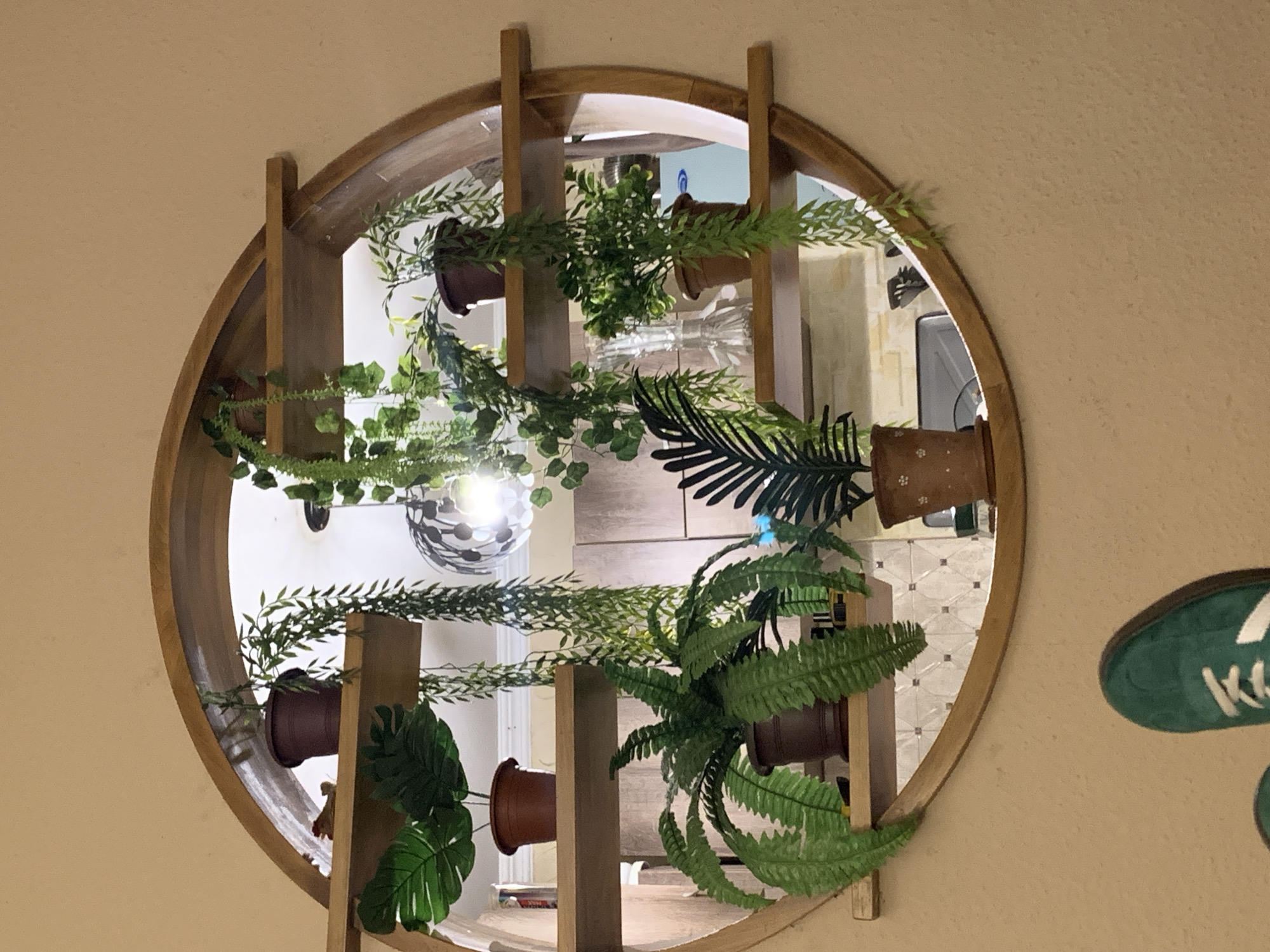 An indoor shot captures a circular wooden shelf mounted on a tan wall, displaying a variety of potted plants. The shelf features three horizontal wooden planks, each holding one or more artificial plants. The leftmost plank on the top shelf holds a dark brown pot with a small, leafy plant, and a miniature decorative object. The middle plank on the top shelf contains a dark brown pot with a trailing vine. Below this, on the second shelf from the top, a large Monstera leaf plant sits in a brown pot on the left. The third shelf down has a large, bushy fern in a brown pot on the left, and a dark-leafed palm-like plant in a brown, white-dotted pot on the right. Cascading vines hang from various points on the shelf. The background visible through the circular shelf shows a tiled wall, a light fixture, and kitchen cabinetry. In the foreground, the tops of two green sneakers are visible, suggesting someone is sitting in front of the shelf.