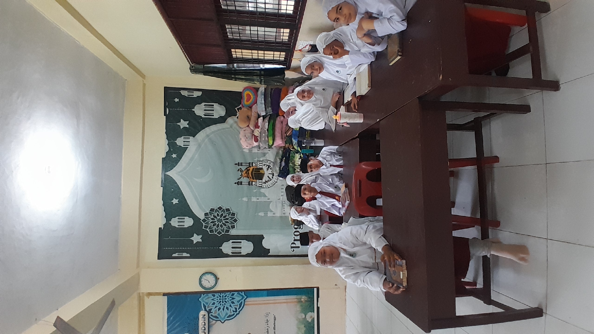 A photograph shows a group of children, dressed in white religious attire, seated at desks in a classroom setting. The location is Lhokseumawe, Indonesia, as indicated by the banner in the background featuring a stylized image of a mosque and Arabic script. The children are arranged on both sides of the tables, which appear to be stained dark wood, accompanied by red chairs. Some children are holding books or are looking towards the camera, while others are engaged in reading. The backdrop displays a large banner with images associated with Islamic culture, and a smaller poster featuring a picture of a blue floral and geometric design hangs below a round clock. A window can be seen in the upper portion of the photograph. The room is illuminated with overhead lighting.