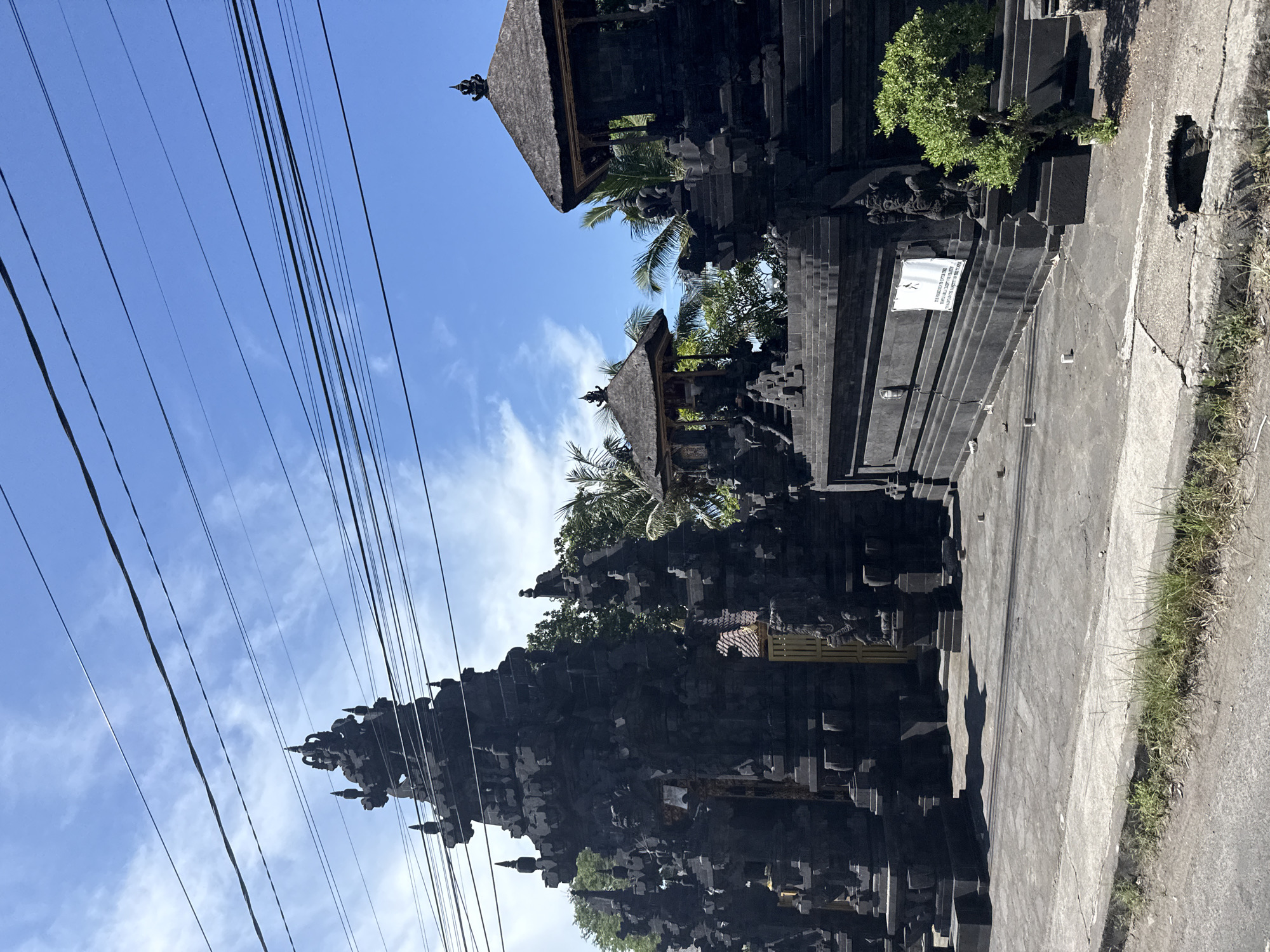 The image captures a street view in Batununggal, Indonesia, showcasing a traditional Balinese temple complex. The temple structures, constructed from dark stone, dominate the scene. The architecture features intricate carvings and tiered roofs. Utility lines stretch across the blue sky with scattered clouds. The foreground includes a concrete pathway or road, with some vegetation along the edge. A small sign is visible on one of the temple walls.