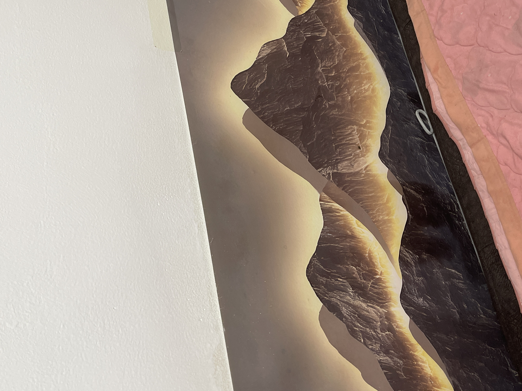 The image depicts a close-up view of a wallpaper featuring a dramatic mountain landscape at sunset. The mountains are rendered with deep shadows and highlights, creating a sense of texture and depth. The sky transitions from a dark, star-speckled expanse at the top to a warm, golden glow behind the peaks, suggesting the sun is setting or rising. The foreground of the image is occupied by a bed with pink and coral colored bedding, indicating this is a domestic setting. A thin, light-colored cable with a white loop is visible on the bedding, partially obscuring the lower portion of the mountain wallpaper. The upper edge of the wallpaper meets a white, textured ceiling. There are no people or animals present in the image.